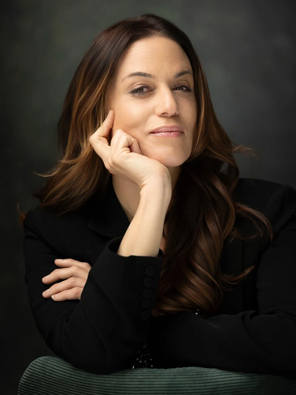 A woman with long, wavy brown hair and a confident expression, resting her chin on her hand, against a dark background.