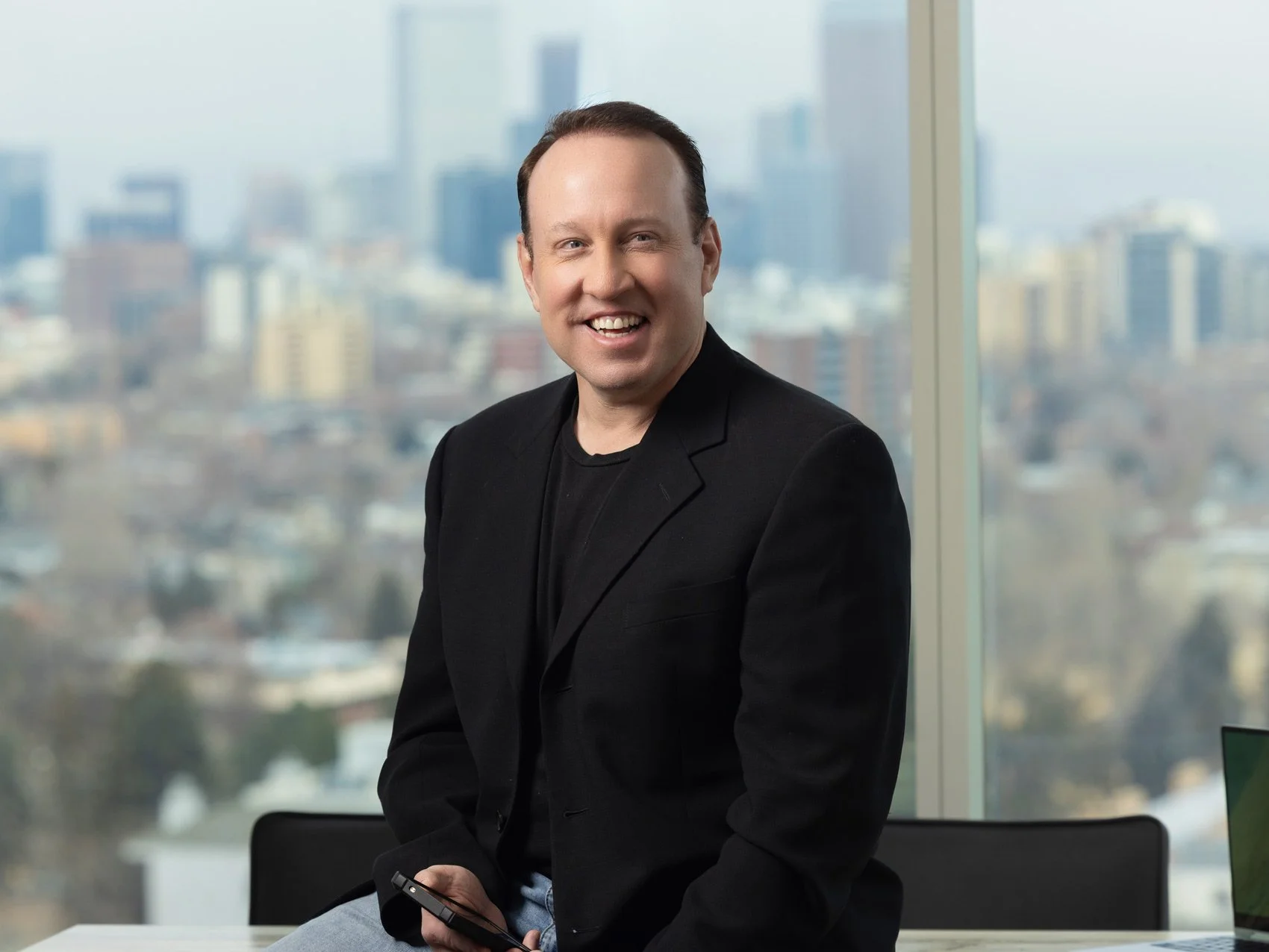 Smiling man in black blazer sitting in an office with a city skyline view behind him, holding a smartphone.