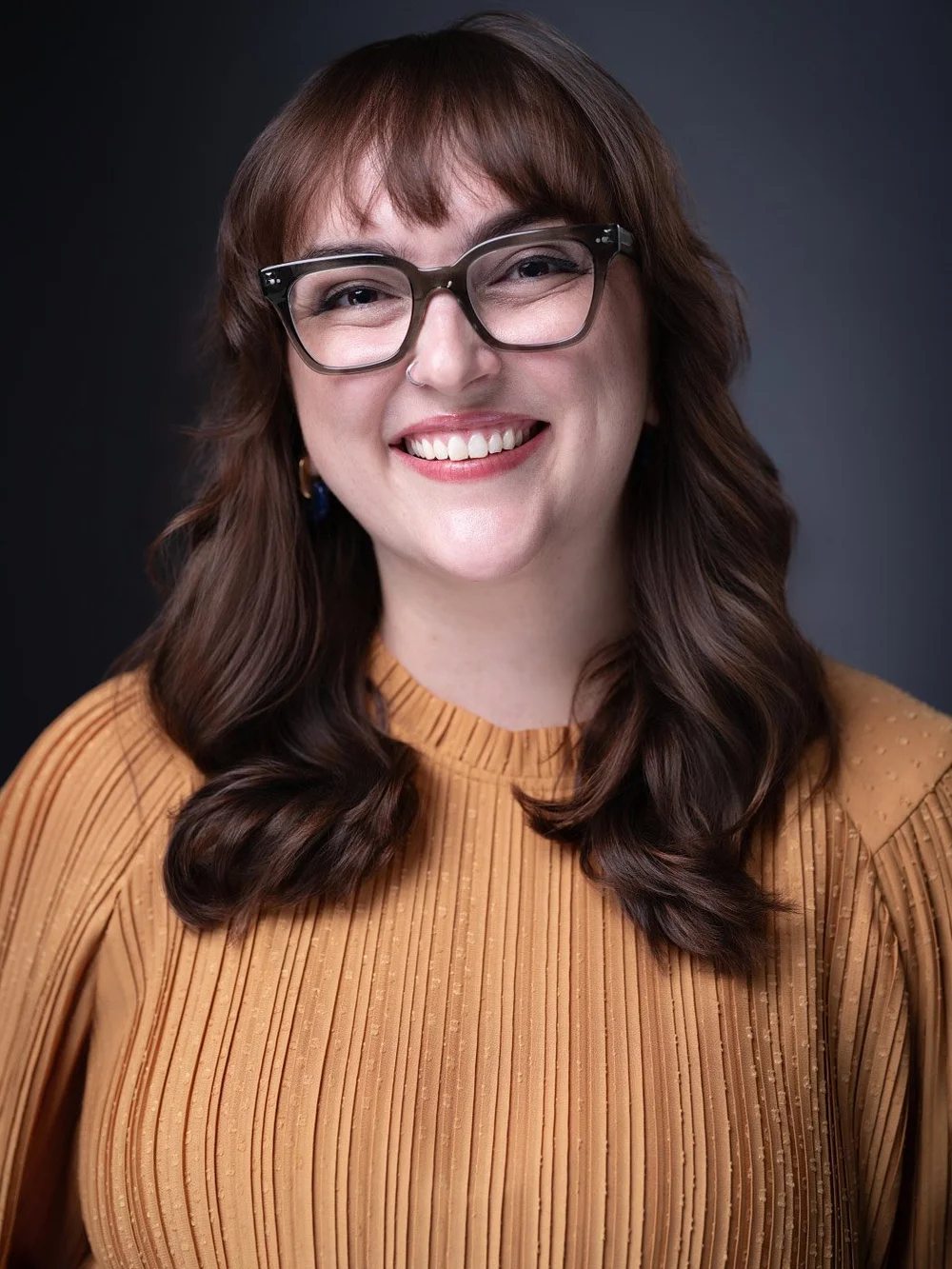 Smiling studio portrait of Kait in glasses and a rust-colored blouse