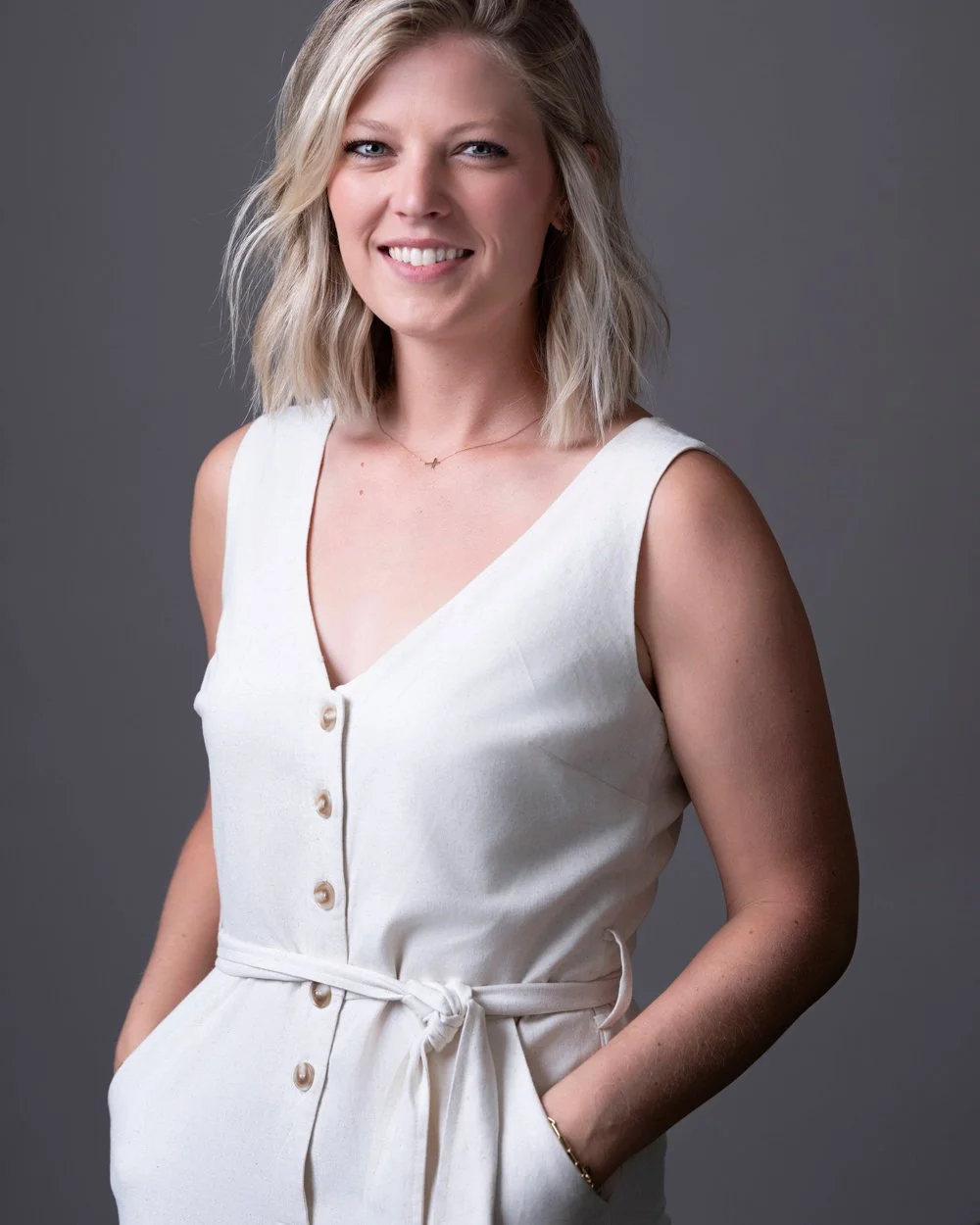 A smiling woman with blonde hair in a sleeveless white dress with buttons and a waist tie, standing against a gray background.