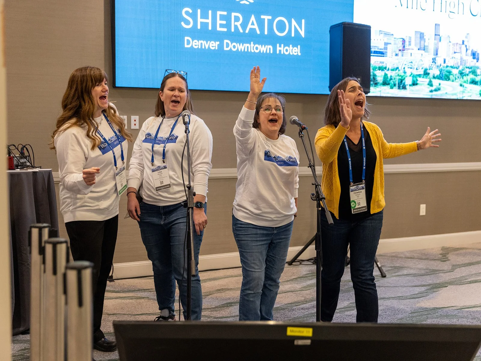 Four women on stage singing into microphones at the Sheraton Denver Downtown Hotel conference.