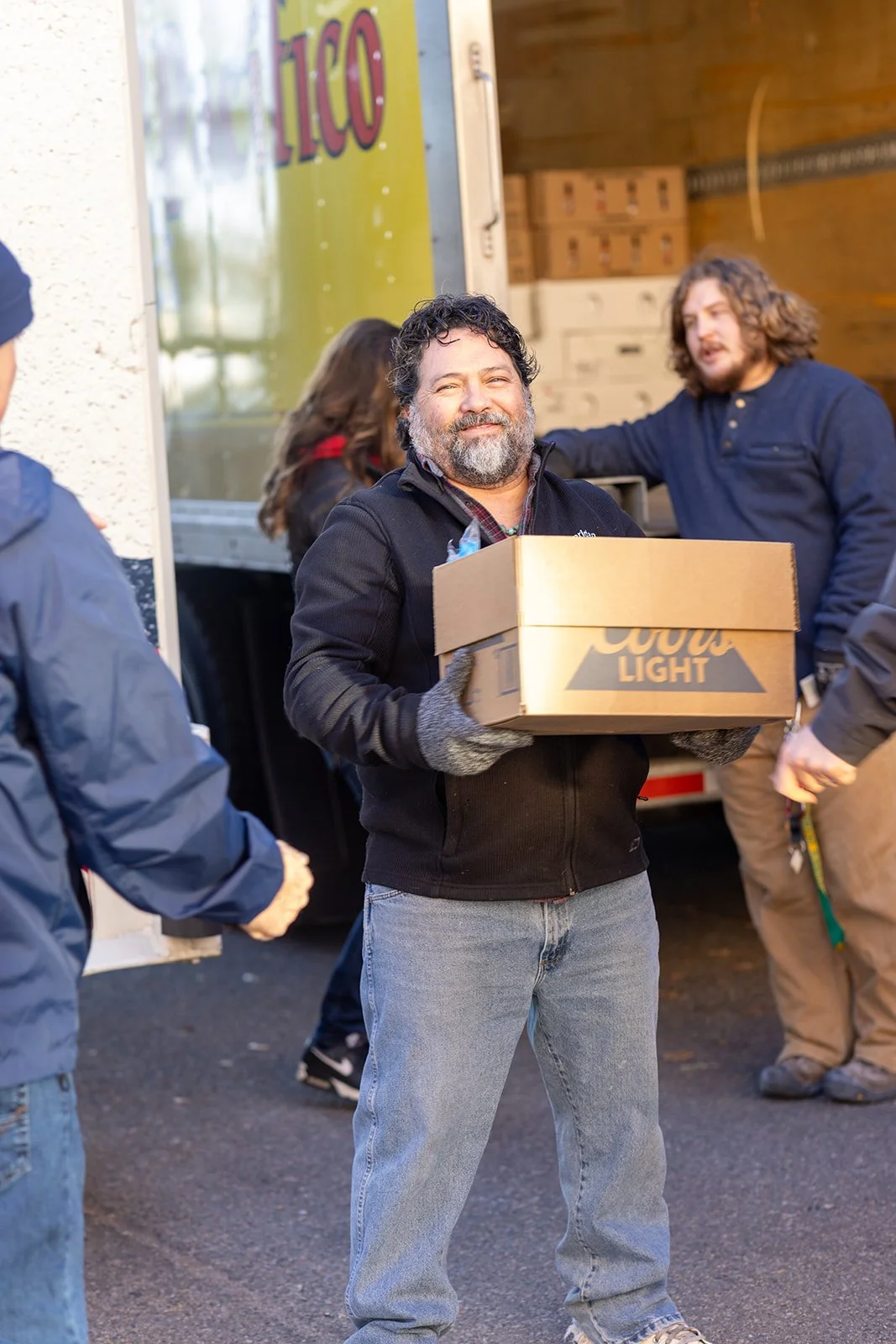 A man with a beard, smiling, holding a box of Coors Light beer, standing outdoors in front of an open delivery truck, with other people around.
