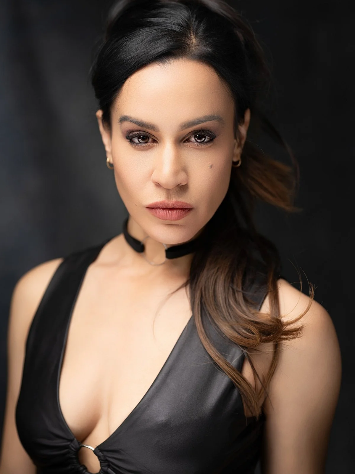 A young woman with dark hair and makeup, wearing a black leather top and choker necklace, looking directly at the camera against a dark background.