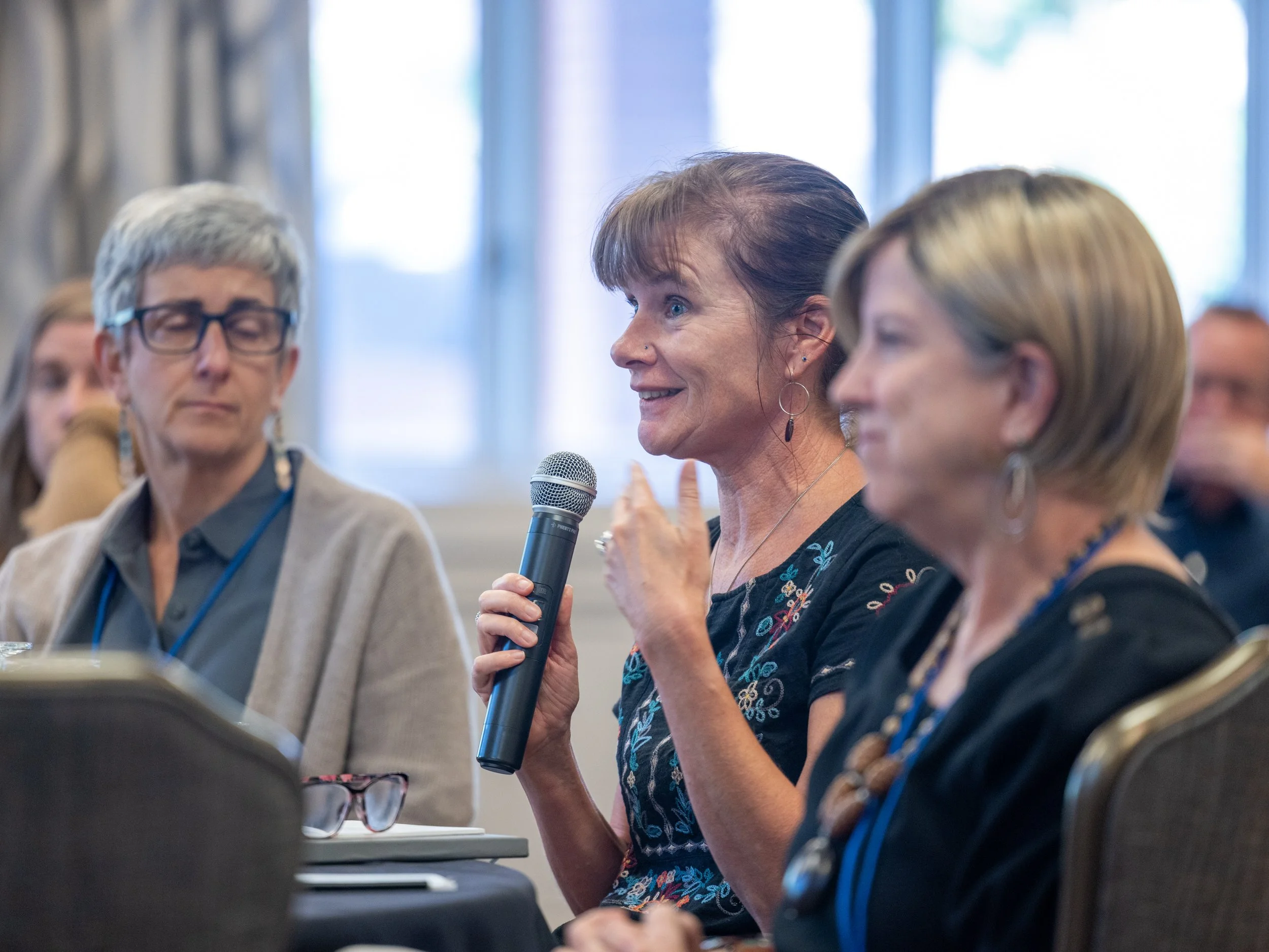 A woman with short brown hair holding a microphone and speaking at a conference, seated next to two other women, one with glasses and short gray hair, and another with short blonde hair, in a bright room with large windows.