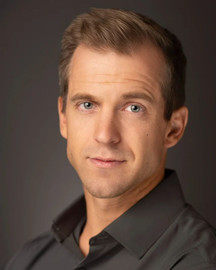 Actor headshot: man in a charcoal button-up shirt with a slight smile against a gray backdrop