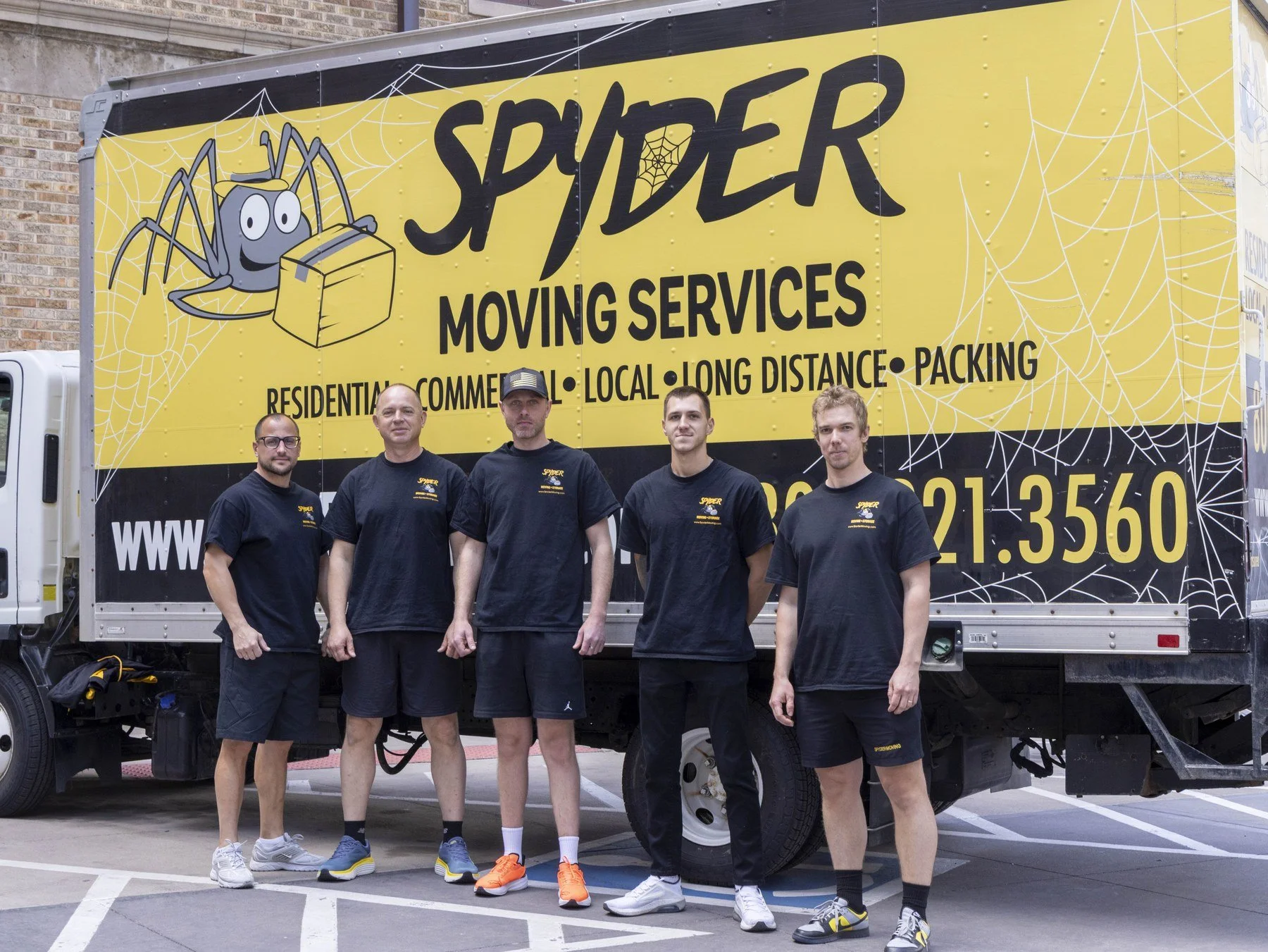 Group of five people standing in front of a yellow moving truck with Spider Moving Services logo and graphic of a spider carrying a box, parked on a city street.