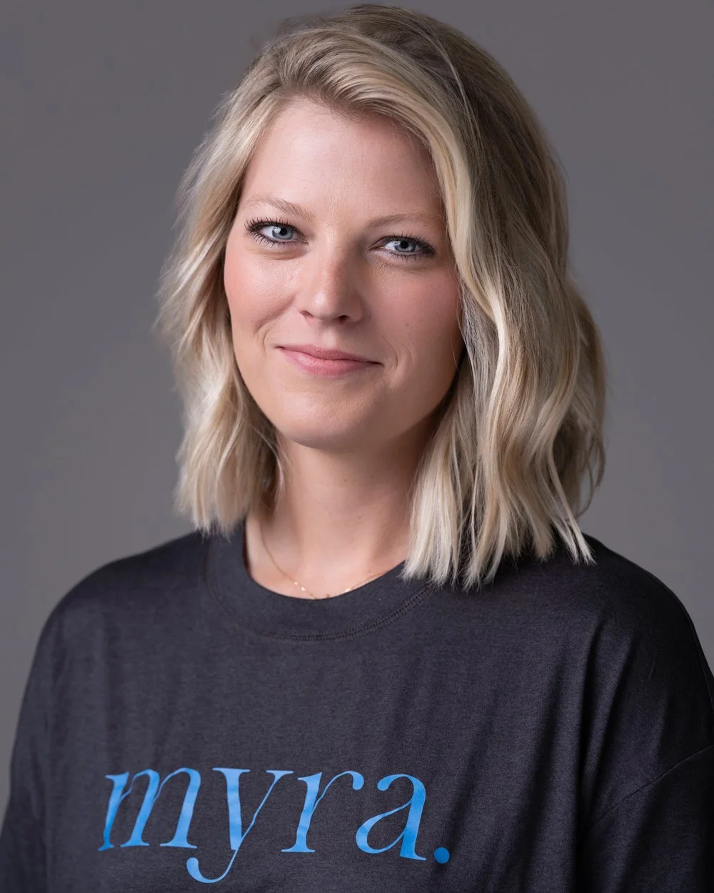 A woman with shoulder-length blonde hair, blue eyes, and light skin, wearing a black T-shirt with the word 'myra.' written on it in blue letters, standing against a plain gray background.