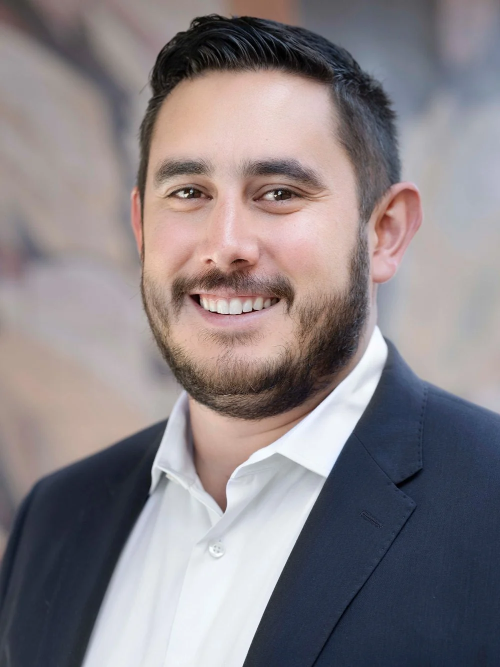 A young man with dark hair, beard, and mustache, wearing a white shirt and dark blazer, smiling for a professional headshot outdoors.