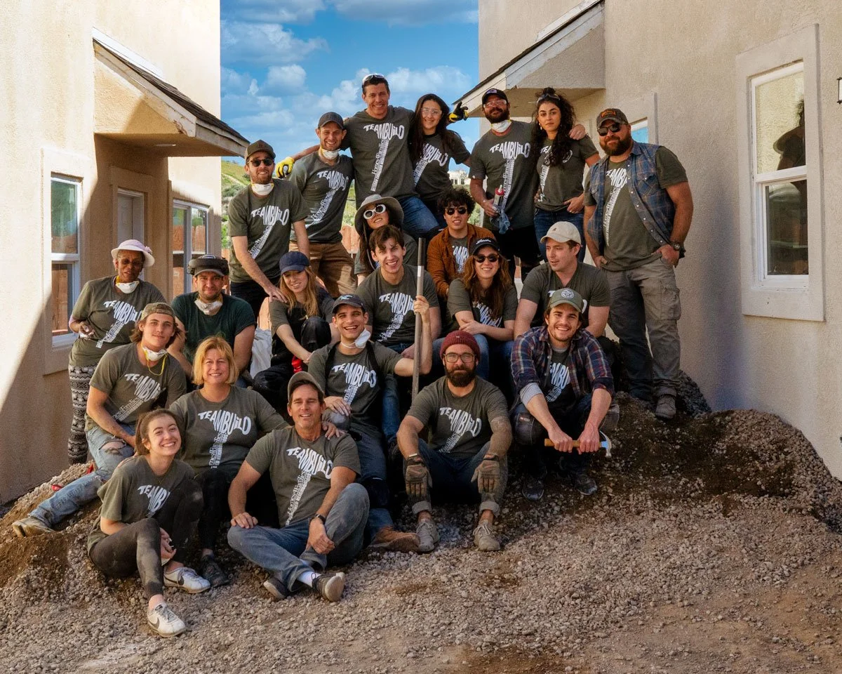 Group of people, wearing matching t-shirts that say "TEAM BUILD," gathered outdoors on a dirt mound building or working on a construction site, with some holding tools, under a partly cloudy sky.