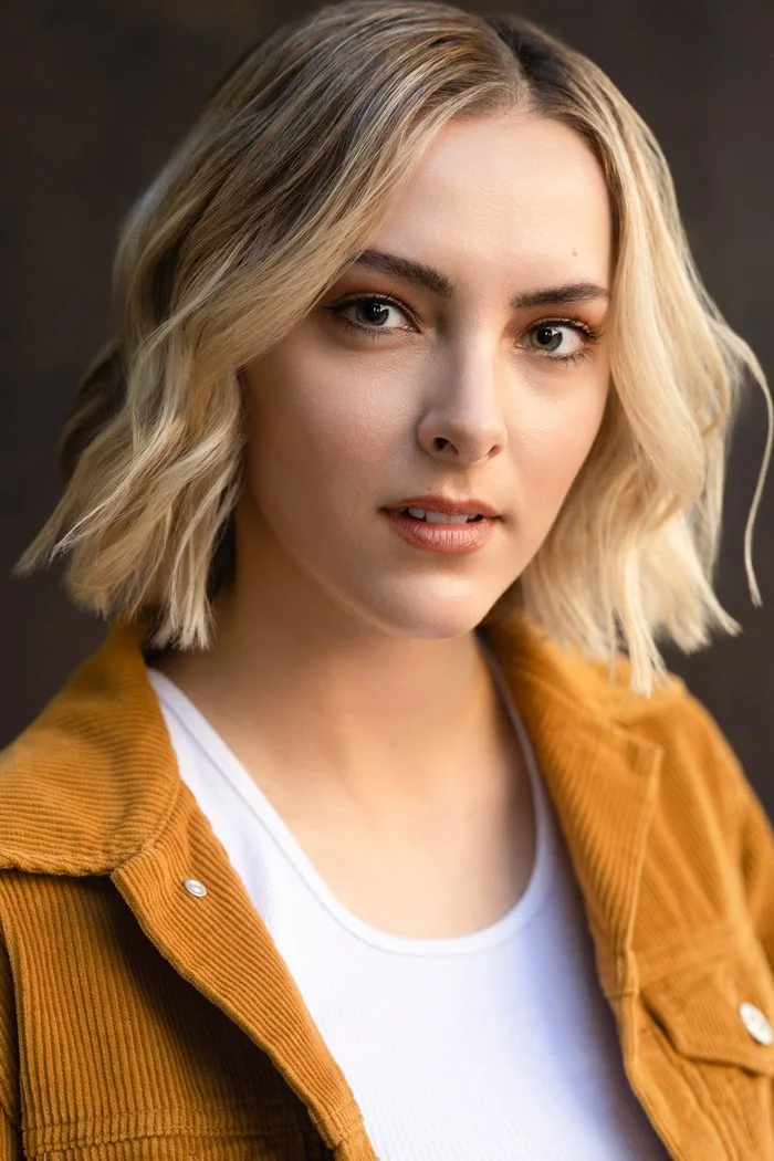 Close-up of a young woman with blonde hair styled in loose waves, wearing a white t-shirt and a mustard-colored corduroy jacket, against a dark background.