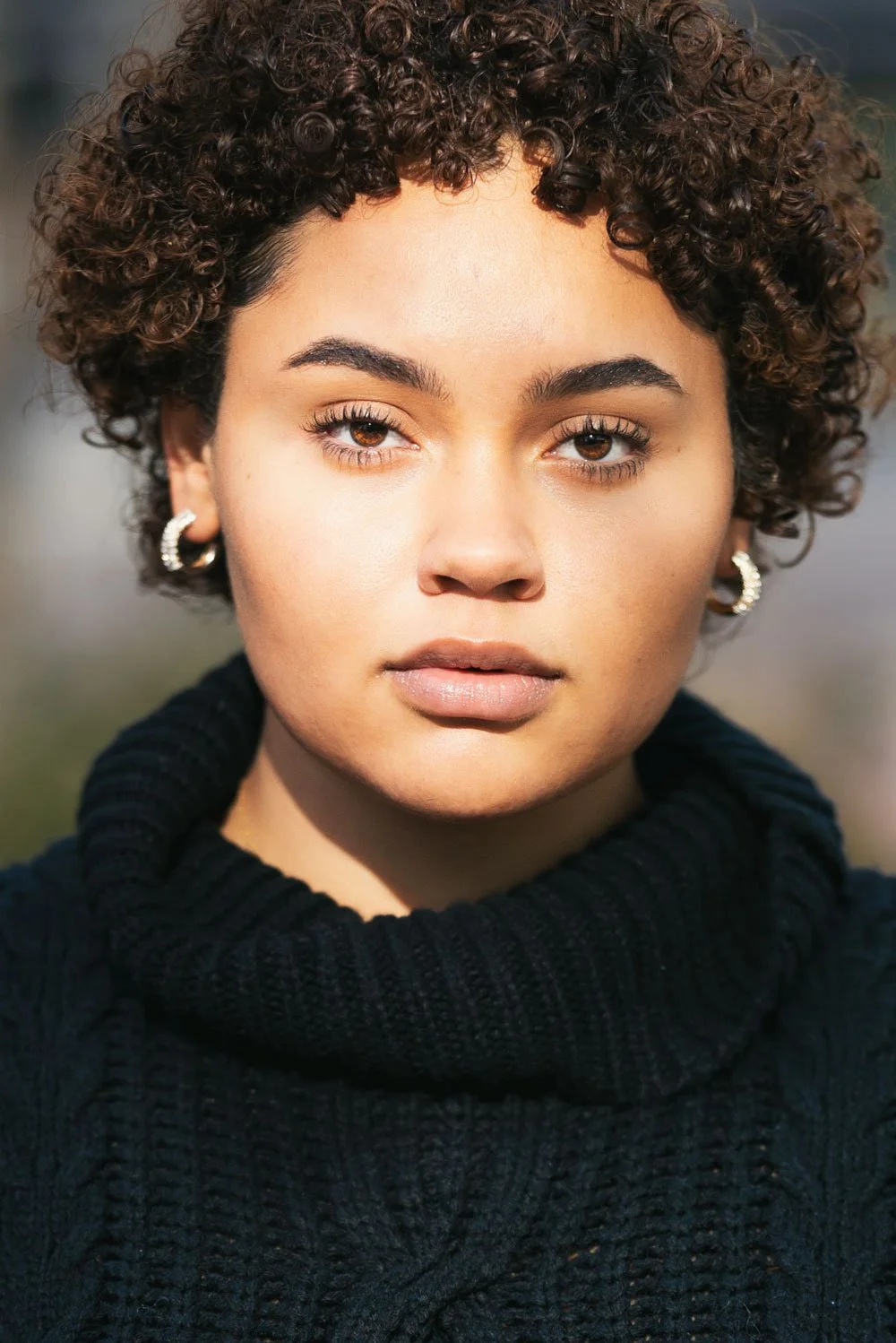 Close-up of a young woman with curly hair, wearing a black turtleneck sweater and hoop earrings, looking directly at the camera with a serious expression.