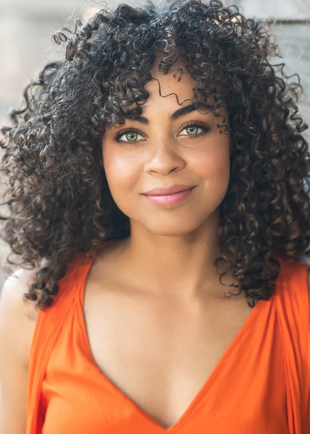 Close-up portrait of a woman with curly dark hair and light skin, wearing an orange top, smiling gently at the camera.