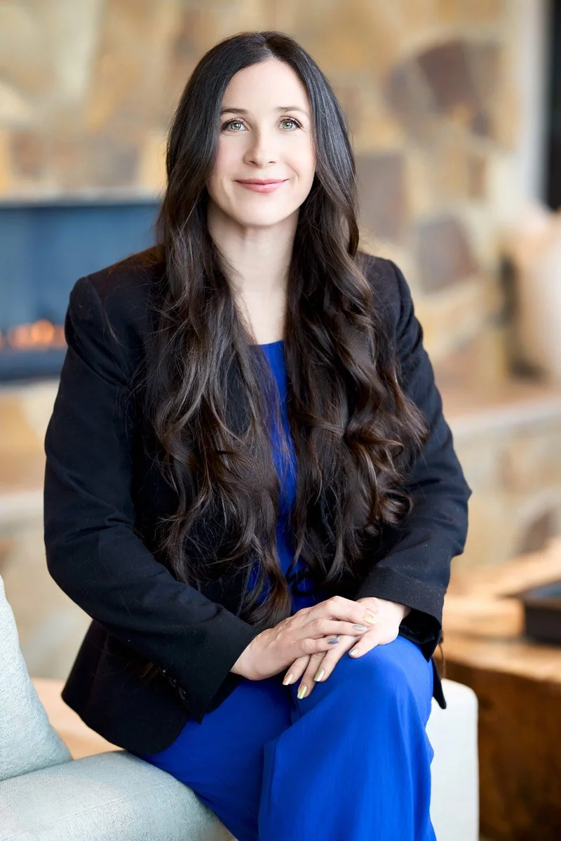 A woman with long dark hair, wearing a black blazer and blue dress, sitting on a light-colored sofa in a cozy, warmly lit room with a stone wall in the background.