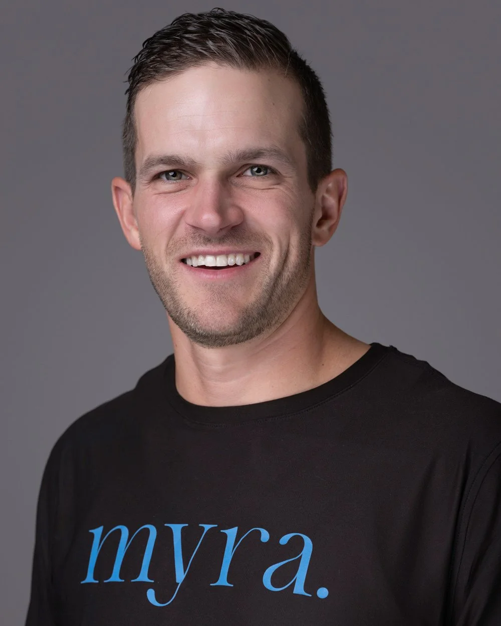 A smiling man with short dark hair and a beard, wearing a black T-shirt with blue text 'myra.' on a gray background.