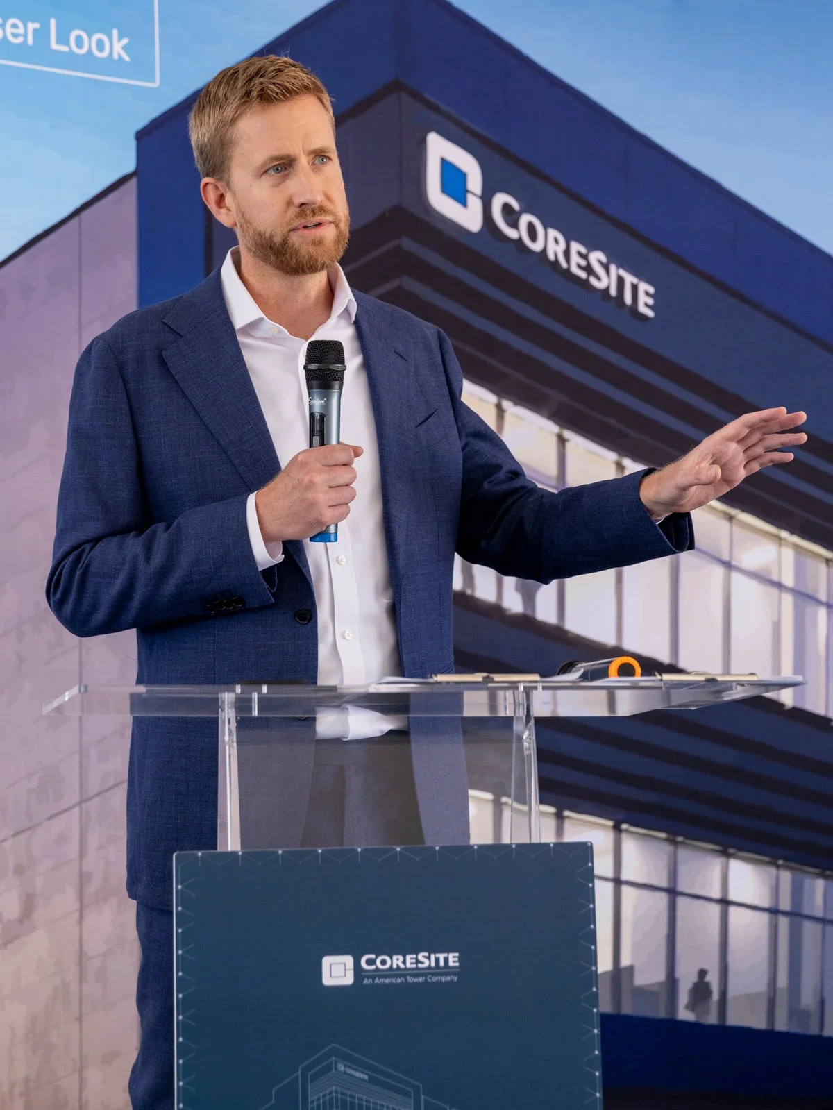 Man in a blue suit giving a presentation at a conference, standing at a podium with a microphone, with a large screen behind him displaying the 'CORE SITE' logo.