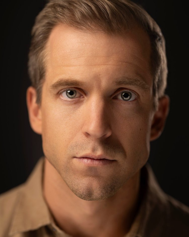 Actor headshot: man in a tan shirt with a direct, steady gaze against a dark backdrop