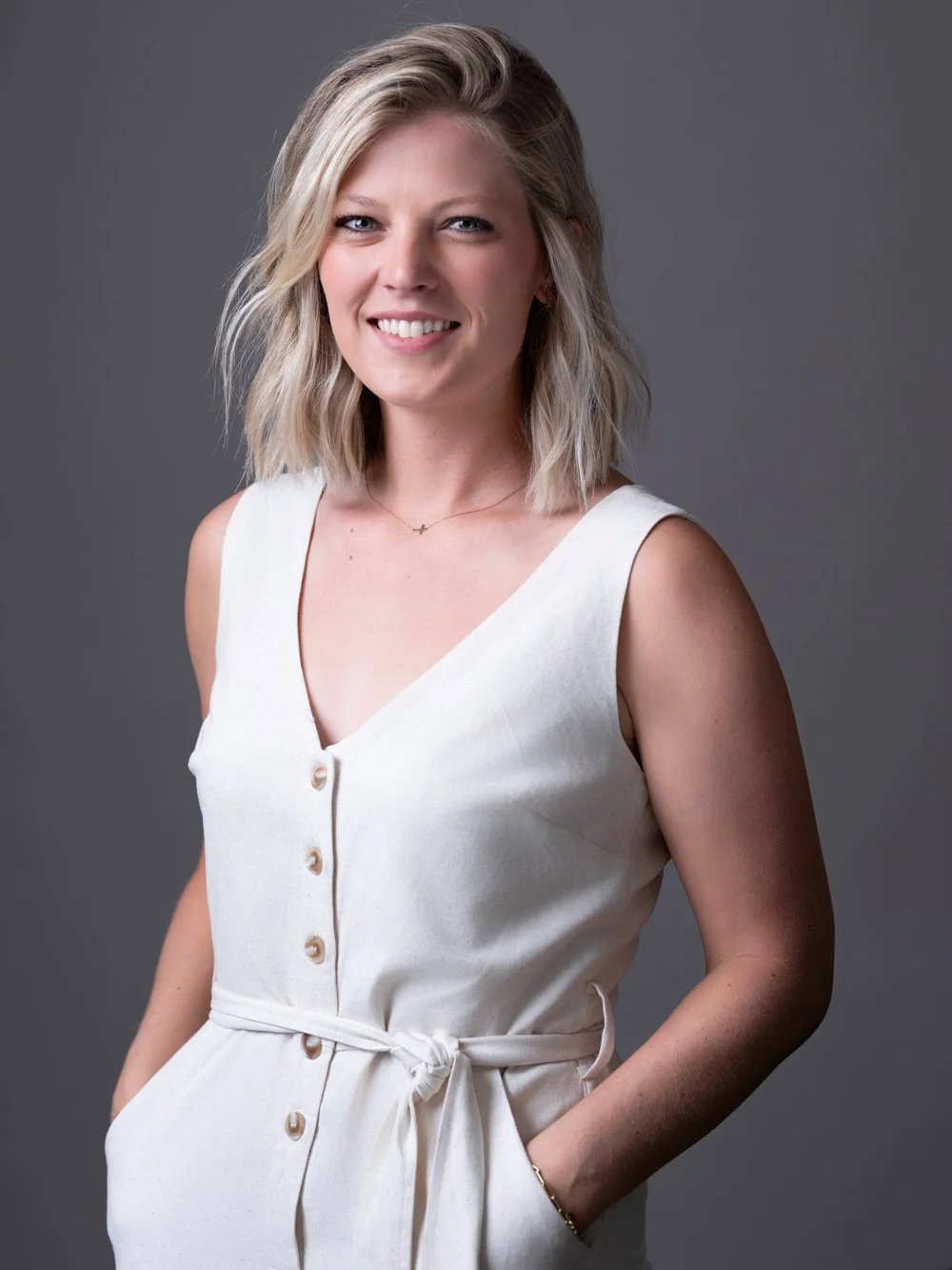 Portrait of a young woman with shoulder-length blonde hair, smiling, wearing a sleeveless white dress with buttons and a tied waist, posing against a gray background.