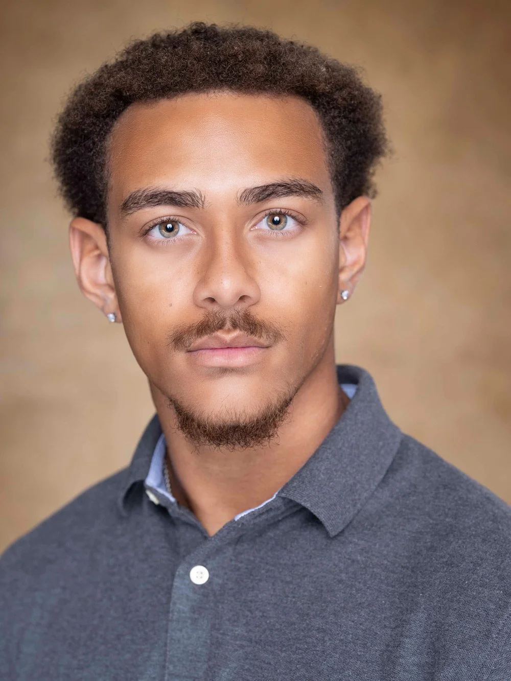 Close-up portrait of a young man with light brown skin, short curly brown hair, blue eyes, a light mustache and goatee, wearing a dark gray collared shirt and small earrings.