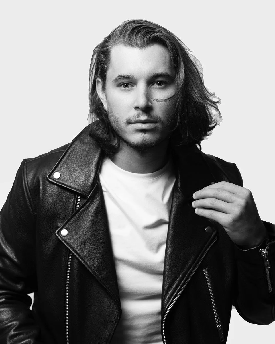 Black and white portrait of a young man with long hair wearing a leather jacket and white T-shirt, looking directly at the camera.