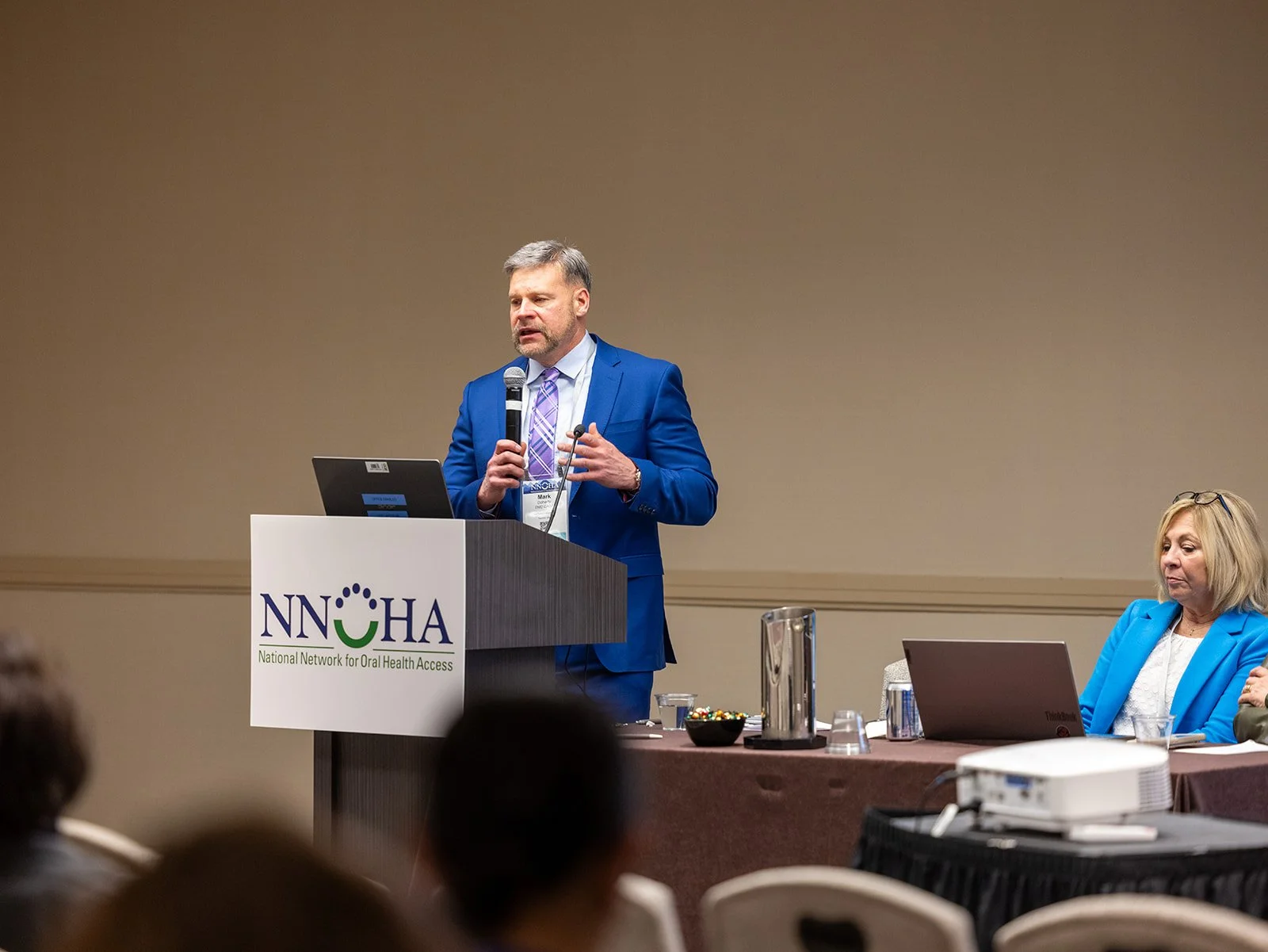 A man in a blue suit speaking at a podium with a laptop, during a conference about oral health. A woman in a blue blazer is seated to his right, with a laptop and glasses on her head, listening.