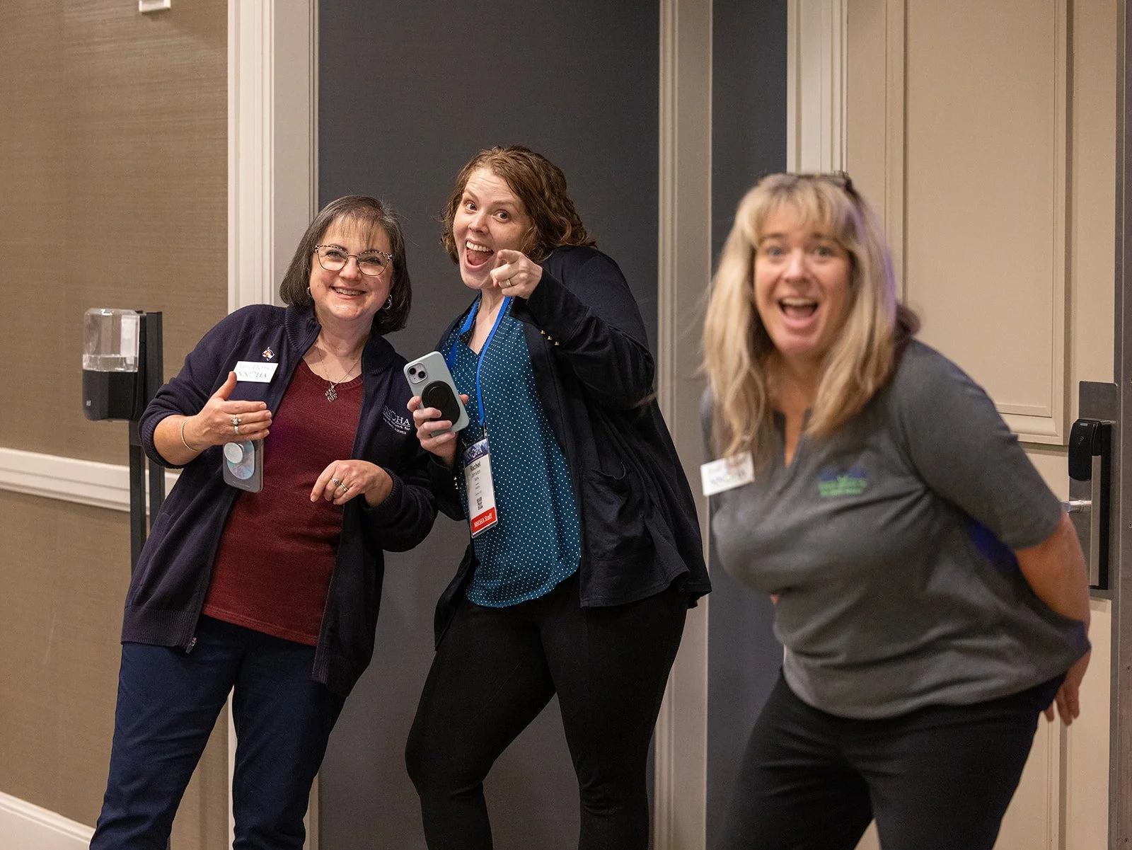 Three women smiling and posing happily at an indoor event, one holding a phone, with a dark door and beige walls in the background.