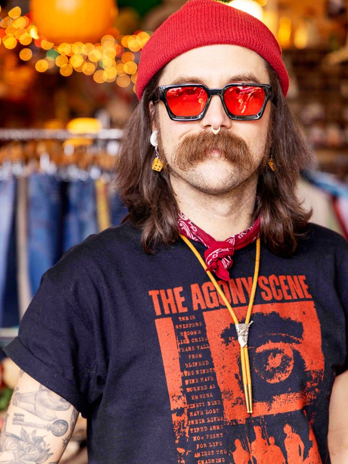 A man with long hair and a mustache wearing a red beanie, red sunglasses, a black T-shirt with red and white graphics, a red bandana around his neck, and earrings, standing in a store with clothing and warm lights in the background.