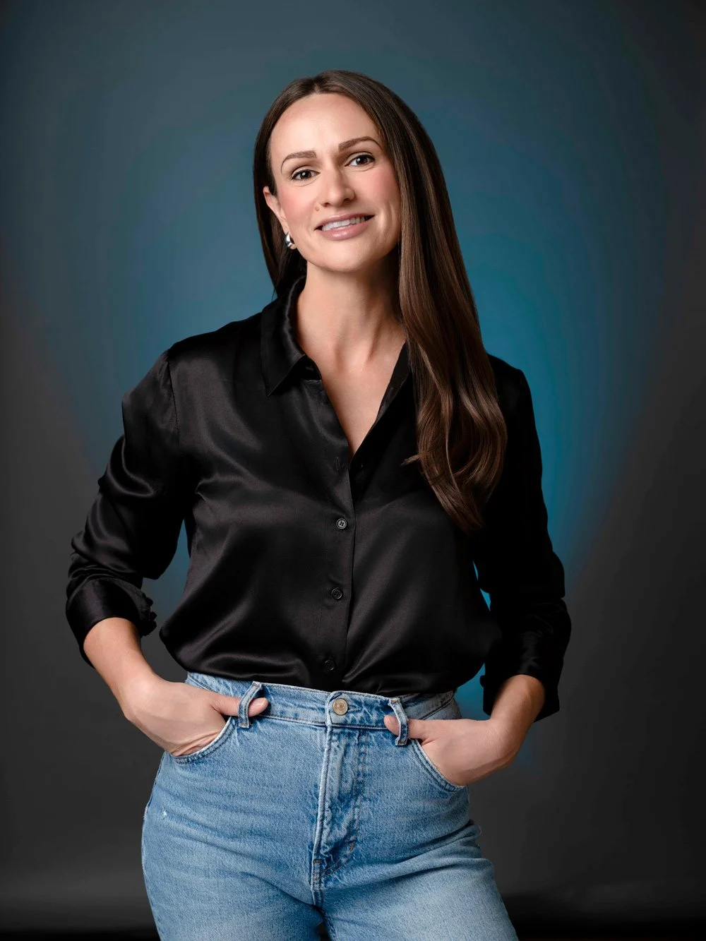 Violet M., who works in the art world, smiling in a black satin shirt and jeans in a Denver studio portrait by Draper Studios