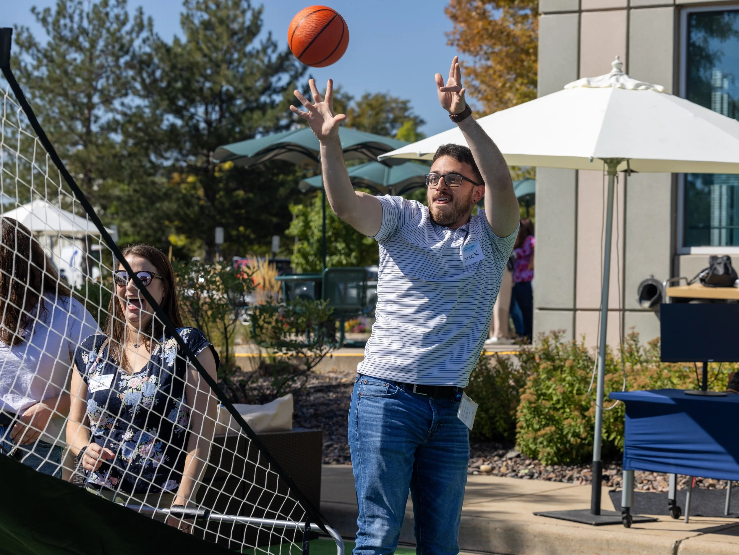 A man playing volleyball outdoors, preparing to hit a volleyball over a net, with people watching and talking nearby under umbrellas on a sunny day.