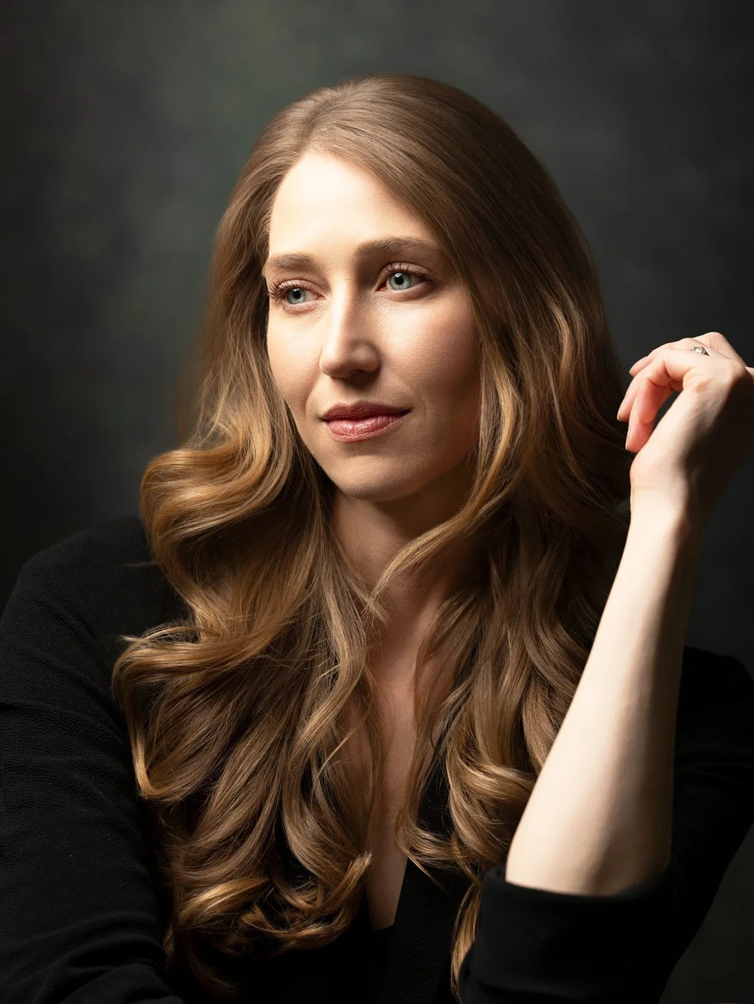 A woman with long, wavy, light brown hair looking thoughtfully to her left against a dark background.