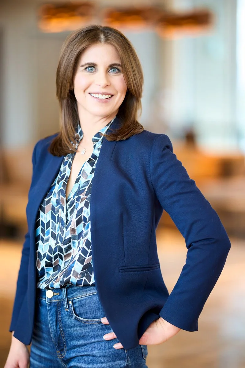 A woman with brown hair and blue eyes, wearing a navy blazer and a patterned blouse, smiling in an indoor setting with warm lighting.
