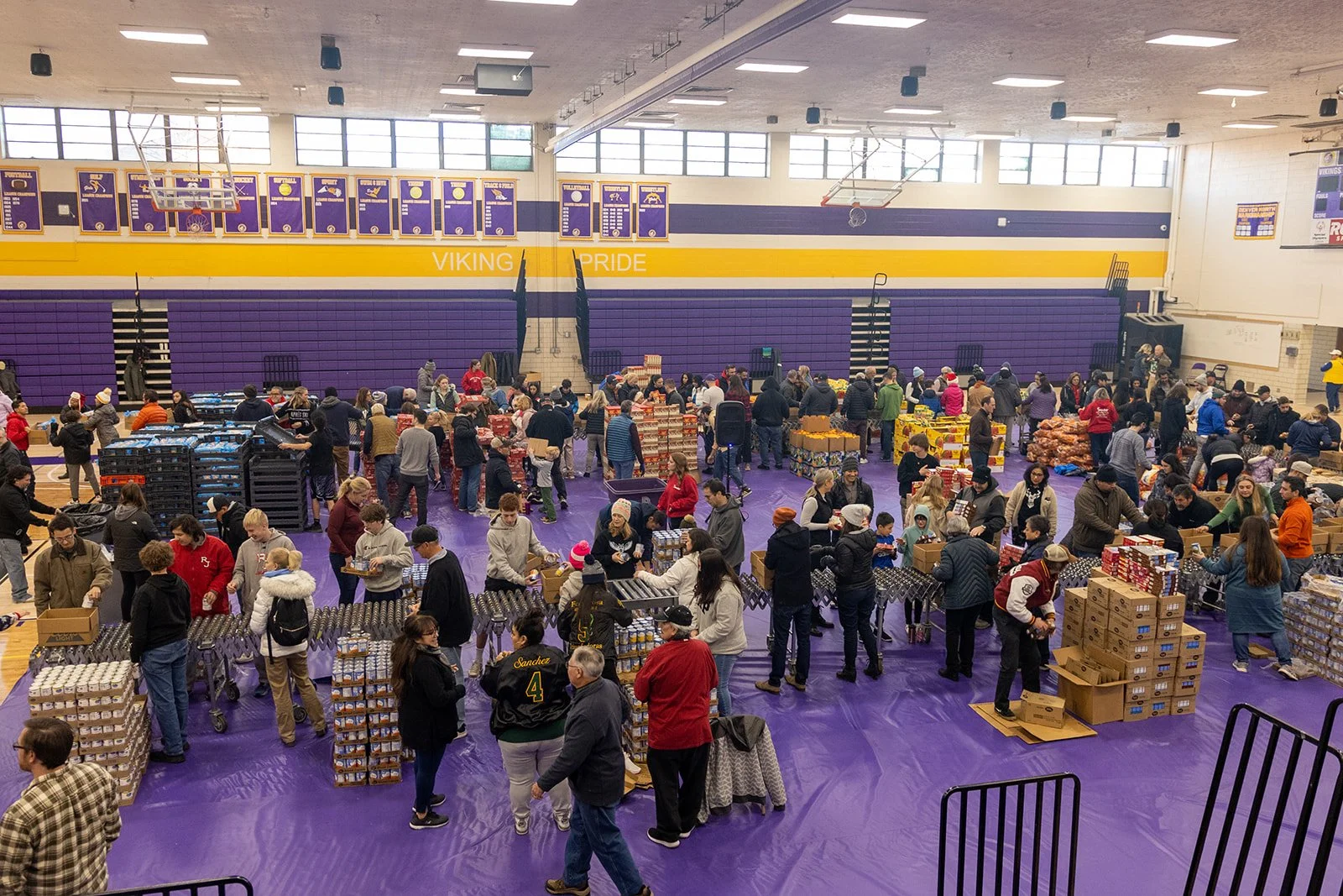 People gathered inside a gymnasium during a food drive event, collecting and organizing boxes of food and supplies. The gym has purple and yellow decor, with banners hanging from the ceiling, and a crowd of volunteers and recipients moving around and