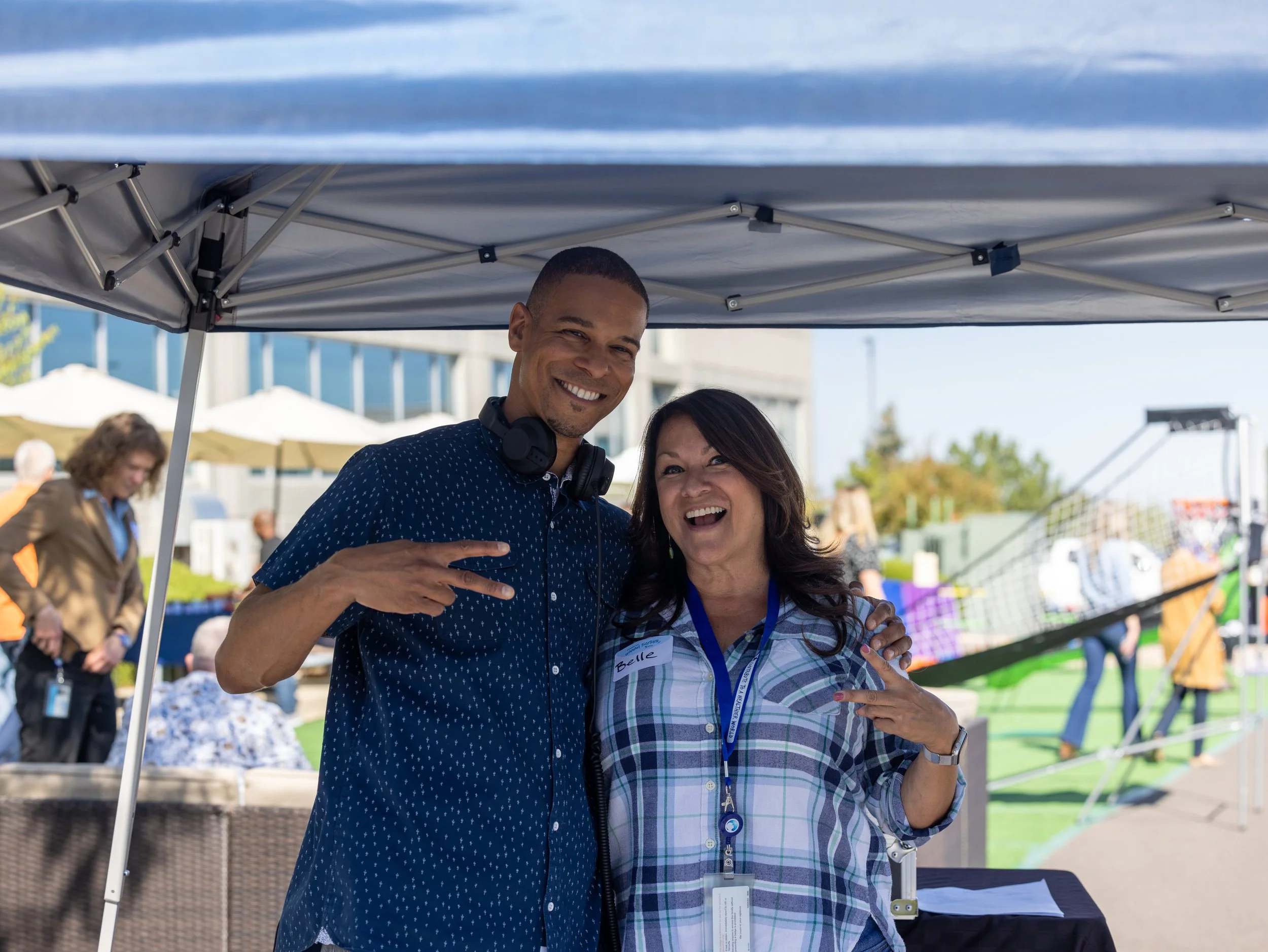 Two smiling people standing under a canopy at an outdoor event, making peace signs with their fingers. The man on the left has short hair, headphones around his neck, and is wearing a dark blue shirt. The woman on the right has long dark hair, a plai