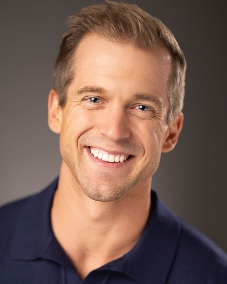 Actor headshot: man in a navy polo with a wide, open smile against a gray backdrop