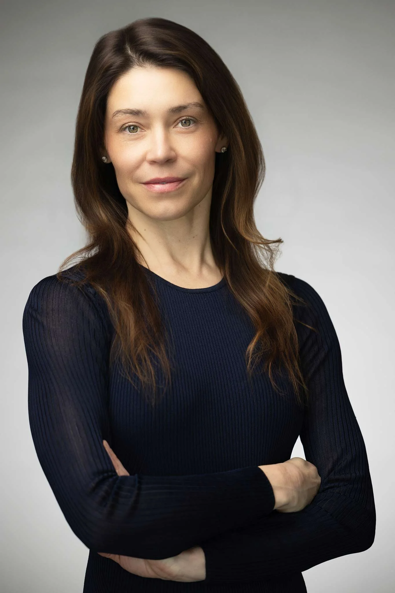 Professional woman with long brown hair, wearing a dark long-sleeve top, standing against a gray background with arms crossed, smiling subtly.