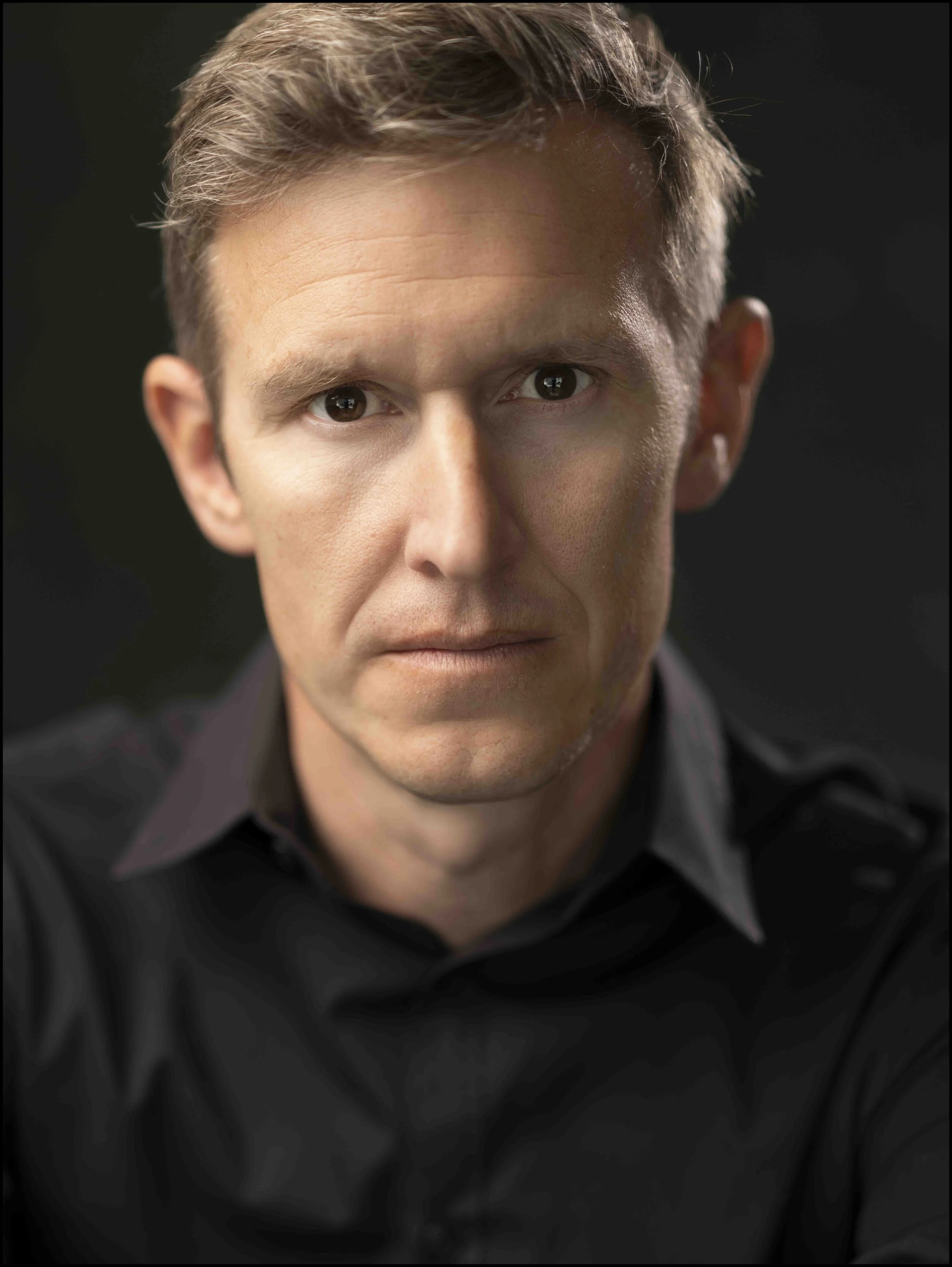 Close-up portrait of a man with short brown hair, wearing a black shirt, against a dark background.