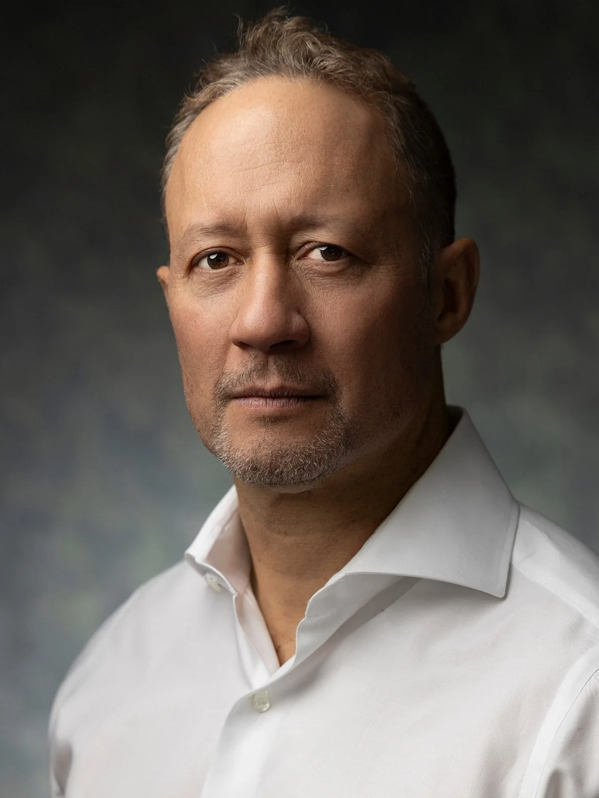Portrait of a middle-aged man with short hair, some gray, wearing a white collared shirt, against a dark, blurred background.