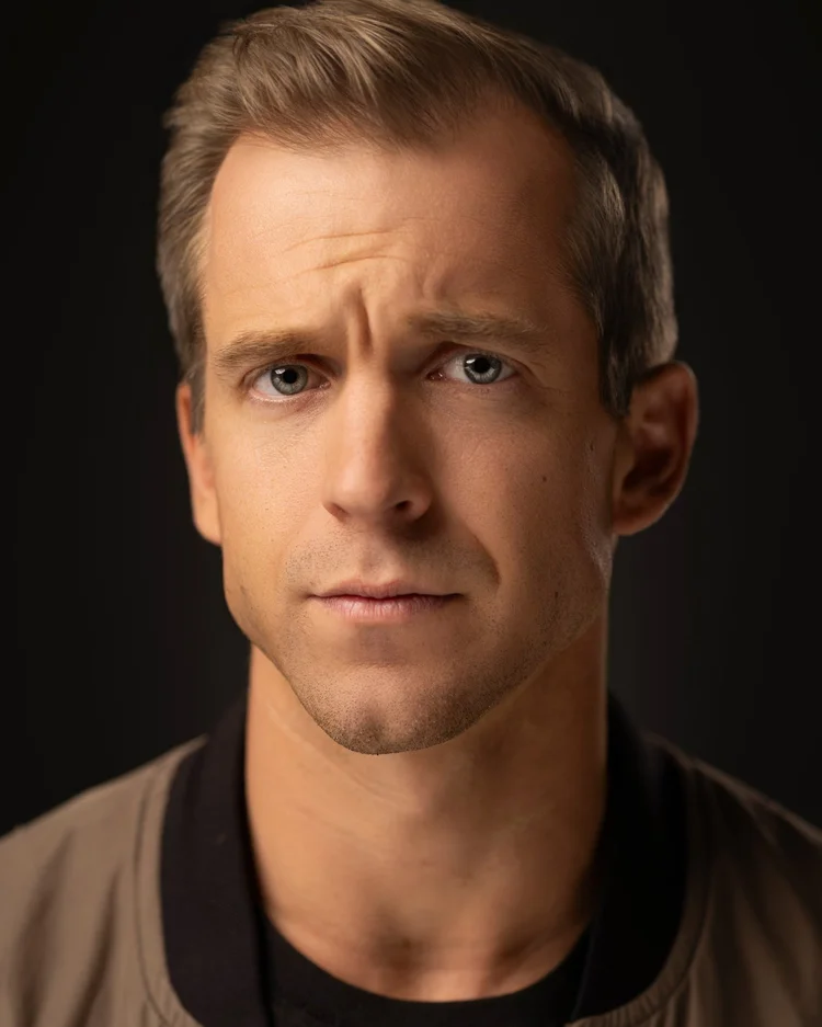 Actor headshot: man in a tan jacket over a black t-shirt, brow furrowed, against a dark backdrop