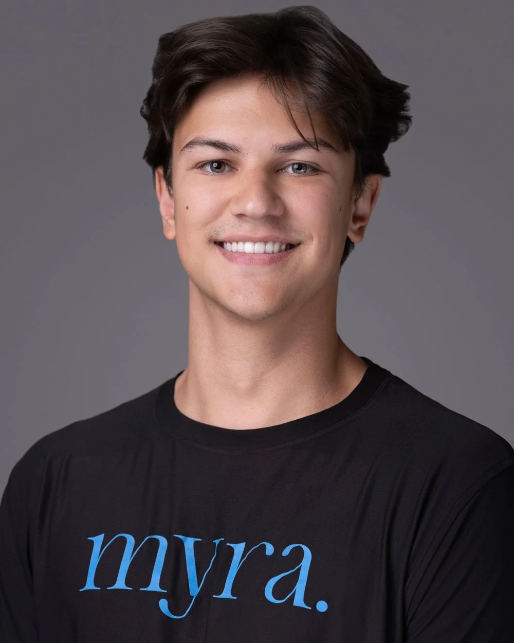 A young man with dark brown hair, light skin, and blue eyes, smiling at the camera. He is wearing a black T-shirt with the word 'myra.' written on it in blue text. The background is a plain gray color.