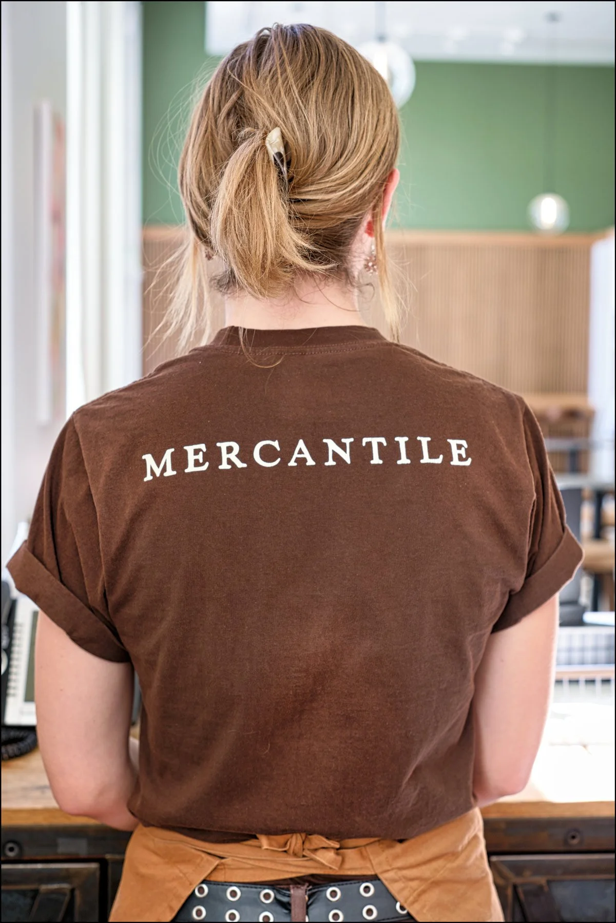Back view of a person with blonde hair tied in a ponytail, wearing a brown t-shirt with the word 'MERCANTILE' printed on the back, standing in what appears to be a cafe or shop.