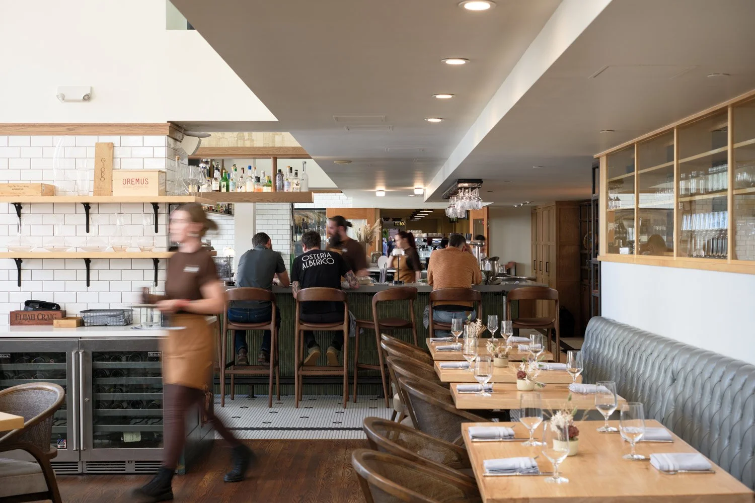 Inside a modern restaurant with a bar area, bar stools, and a seating area with a long table set with wine glasses and small flower arrangements.