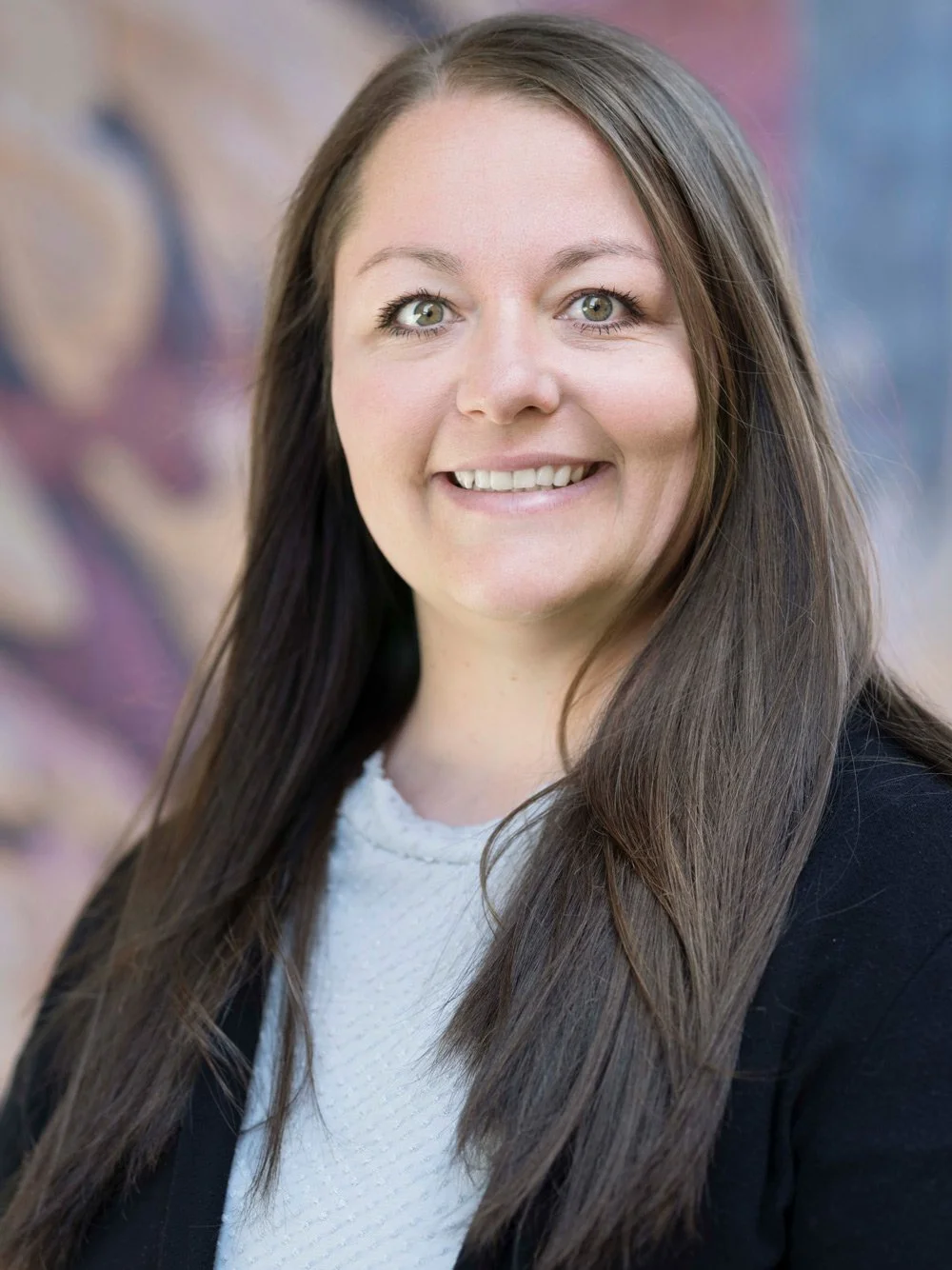 Professional woman with long brown hair smiling, wearing a black blazer and white top, with a blurred colorful background.