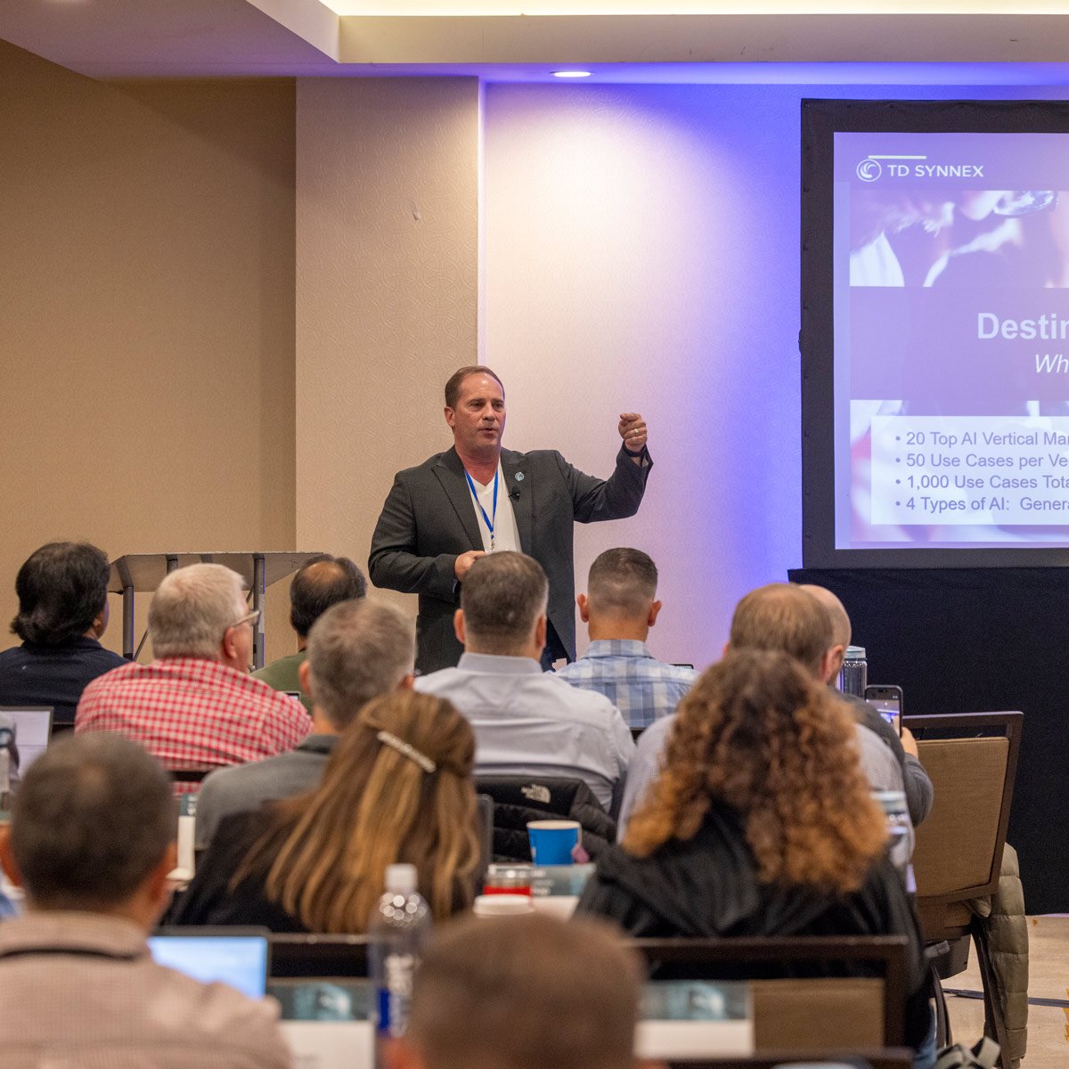 A man in a suit giving a presentation to a seated audience in a conference room, with a large screen displaying slides.