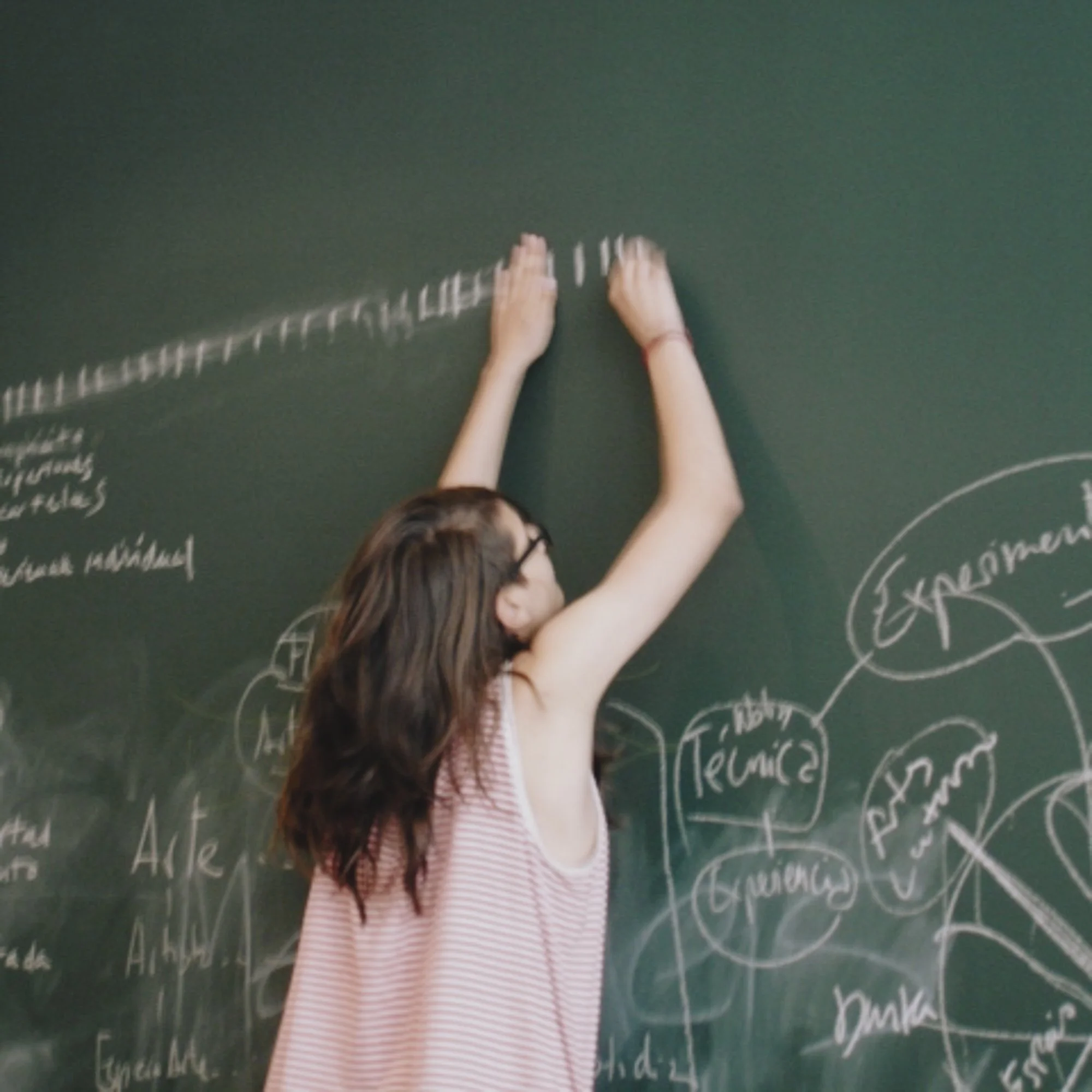 Estudiante con gafas escribe en el pizarrón verde lleno de diagramas y notas en tiza blanca.