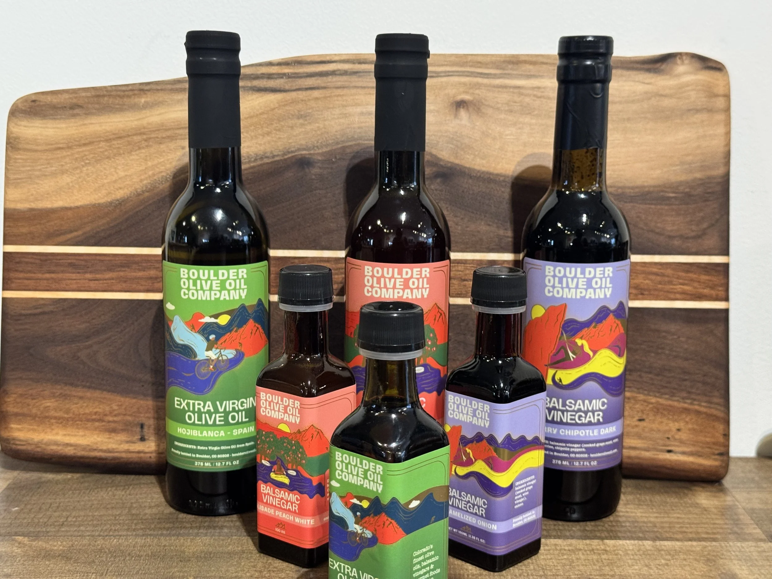 Bottles of Boulder Olive Oil Company variety oils, including extra virgin olive oil, balsamic vinegar, and balsamic vinegar with blueberry chipotle dark flavor, arranged on a wooden display.