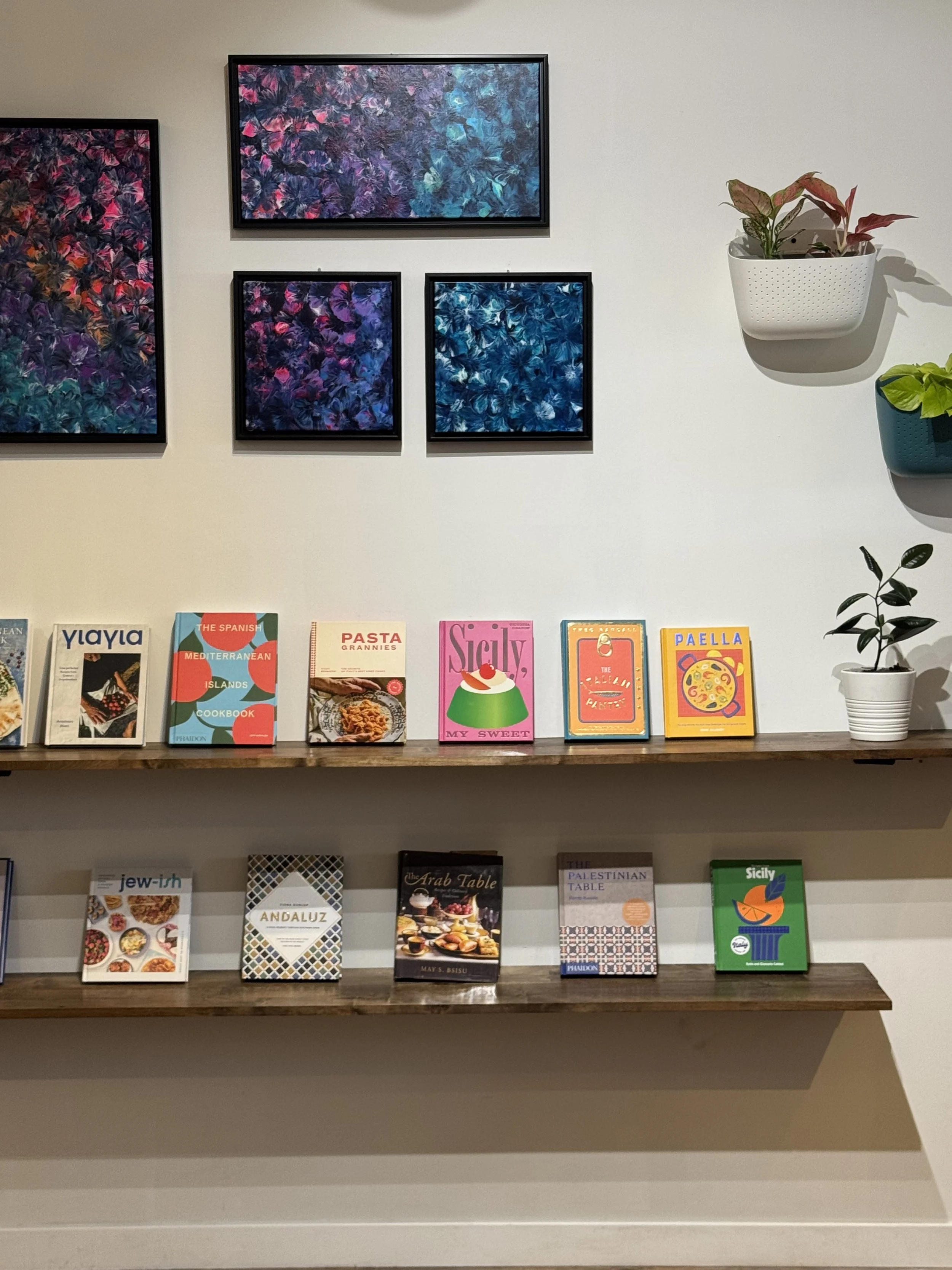 Two wooden shelves display a collection of colorful cookbooks, with three framed abstract paintings hung on a white wall above. Potted plants are positioned on the right side of the shelves.