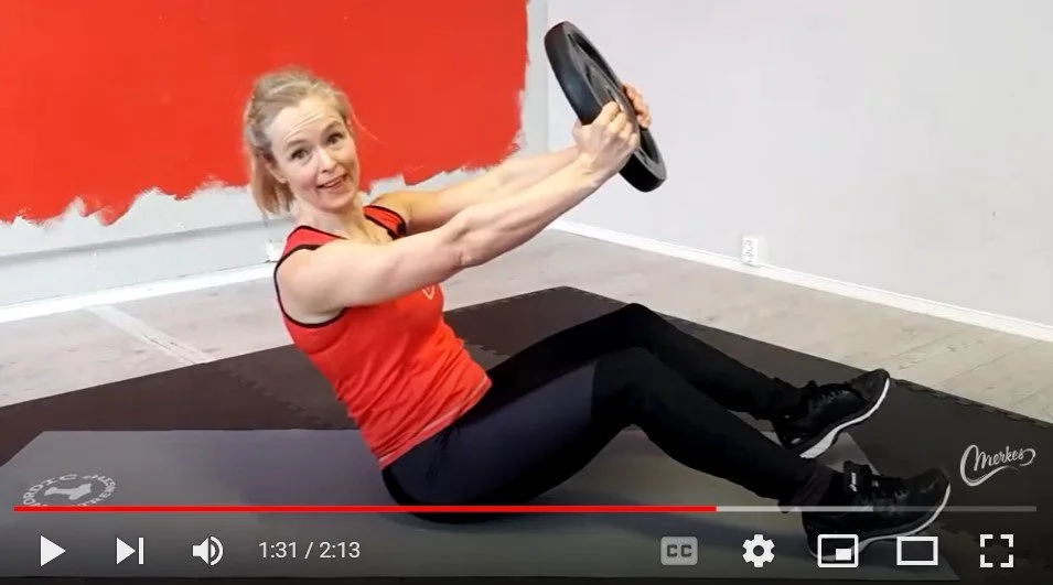 Kvinne i treningsklær utfører mageøvelse på matte, holder en vektplate og har et smilende uttrykk. Bak henne er en vegg delvis malt i rødt. Videoavspillingslinje er synlig nederst.