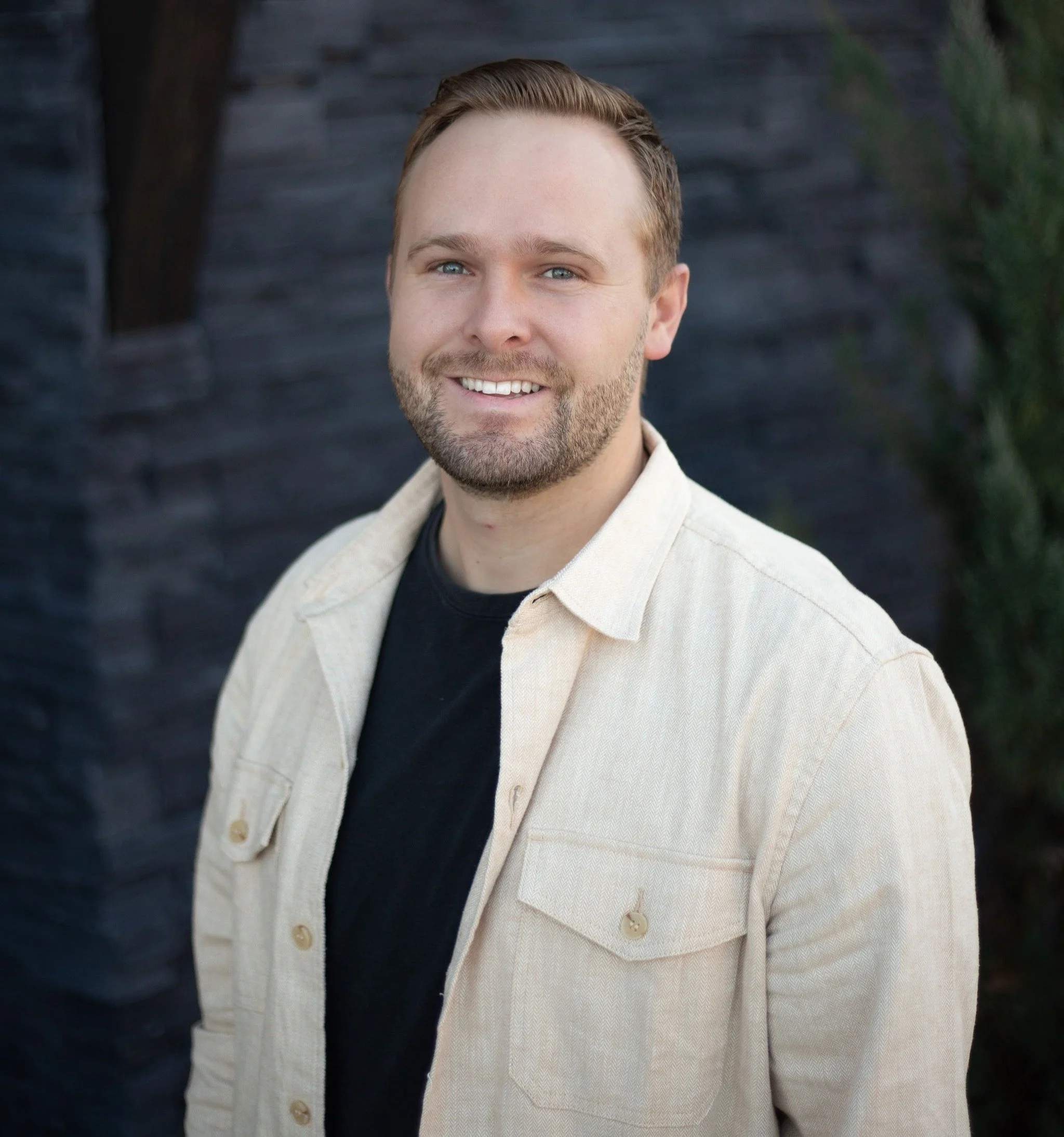 A smiling man with short hair and a beard, wearing a beige jacket over a black shirt, standing outdoors near a dark stone wall with greenery in the background.