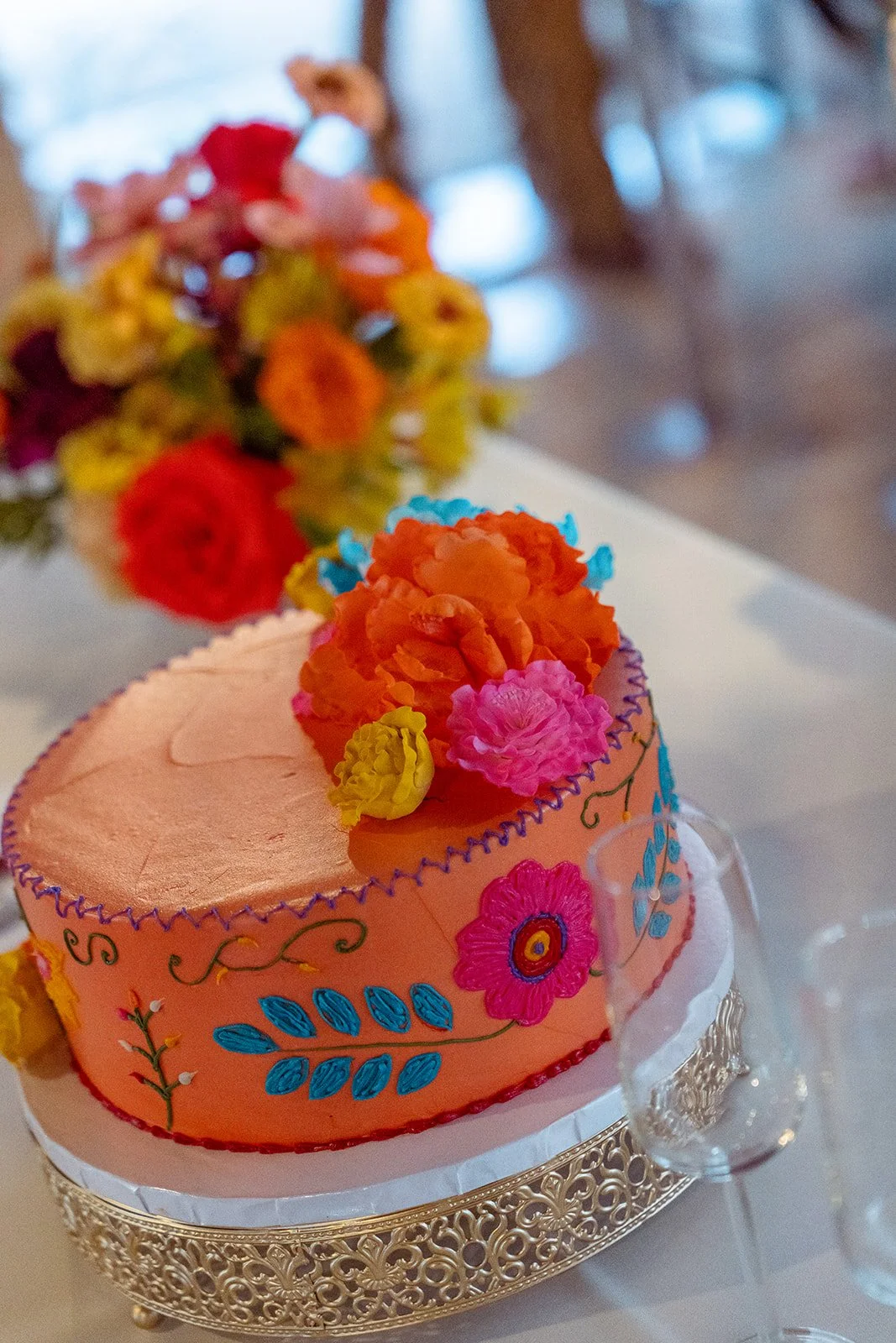 Colorful, floral decorated birthday cake with pink frosting, orange, yellow, pink, and blue flowers on top, presented on a silver ornate cake stand.