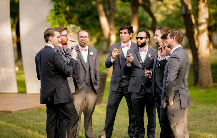 Group of men in suits with bow ties and cigars, standing outdoors on grass with trees, celebrating at a formal event.