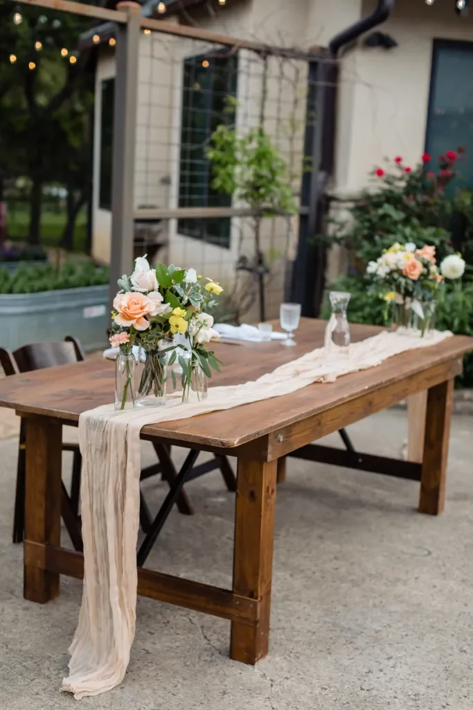 A wooden table set outdoors with floral centerpieces and tableware, decorated with cream-colored fabric runners.